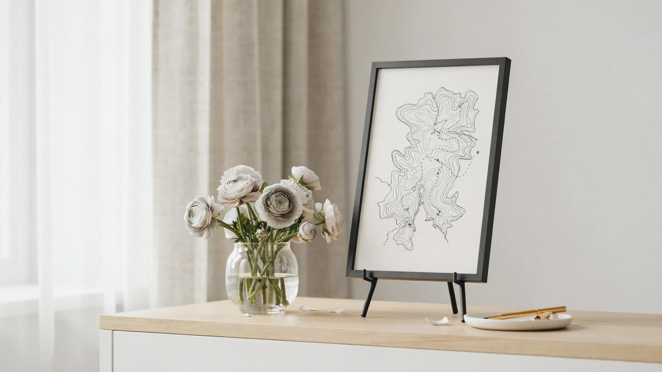 IKEA RIBBA black picture frame on a tabletop easel displaying a minimalist topographic map line art print, styled as a unique creative guest book alternative next to white ranunculus flowers.
