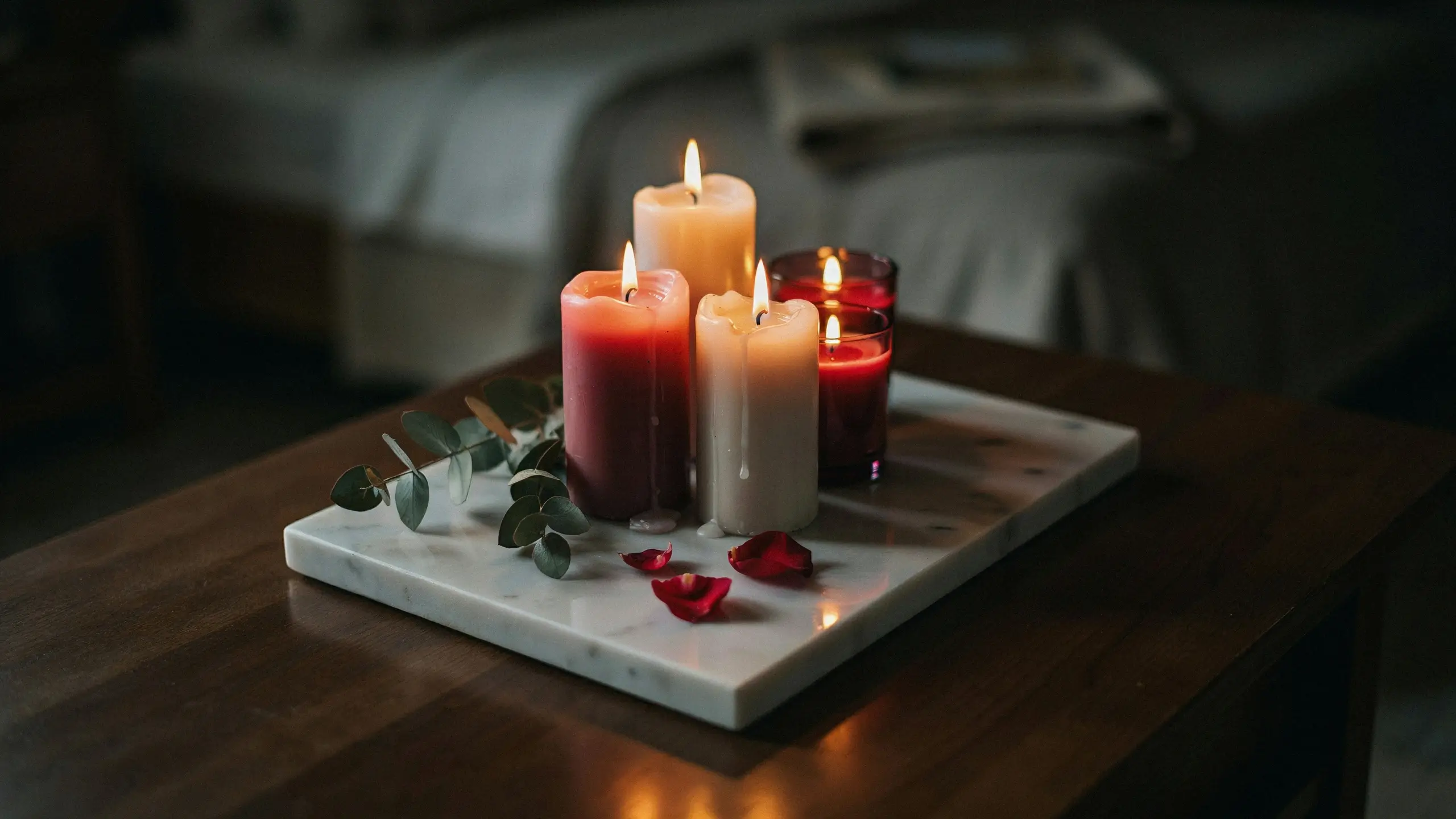 A romantic cluster of red and white pillar candles on a white marble tray with scattered red rose petals and eucalyptus leaves on a dark wooden coffee table in a dimly lit bedroom.