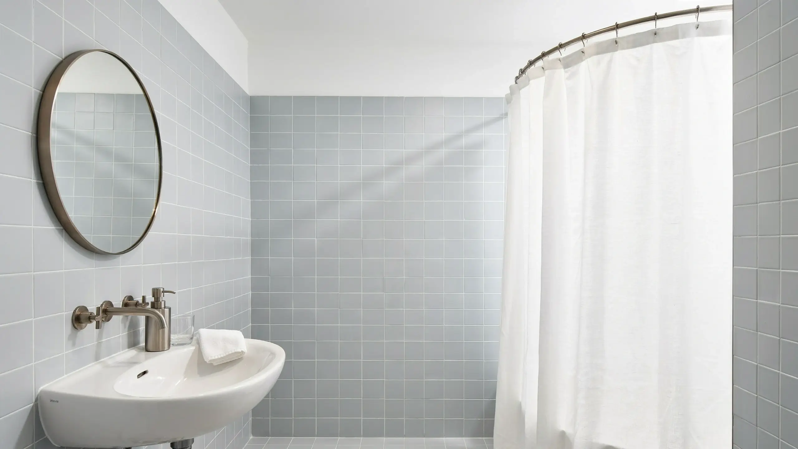 Round wall-mounted mirror and curved shower rod used as rounded fixtures to soften corners in a light blue minimalist bathroom.