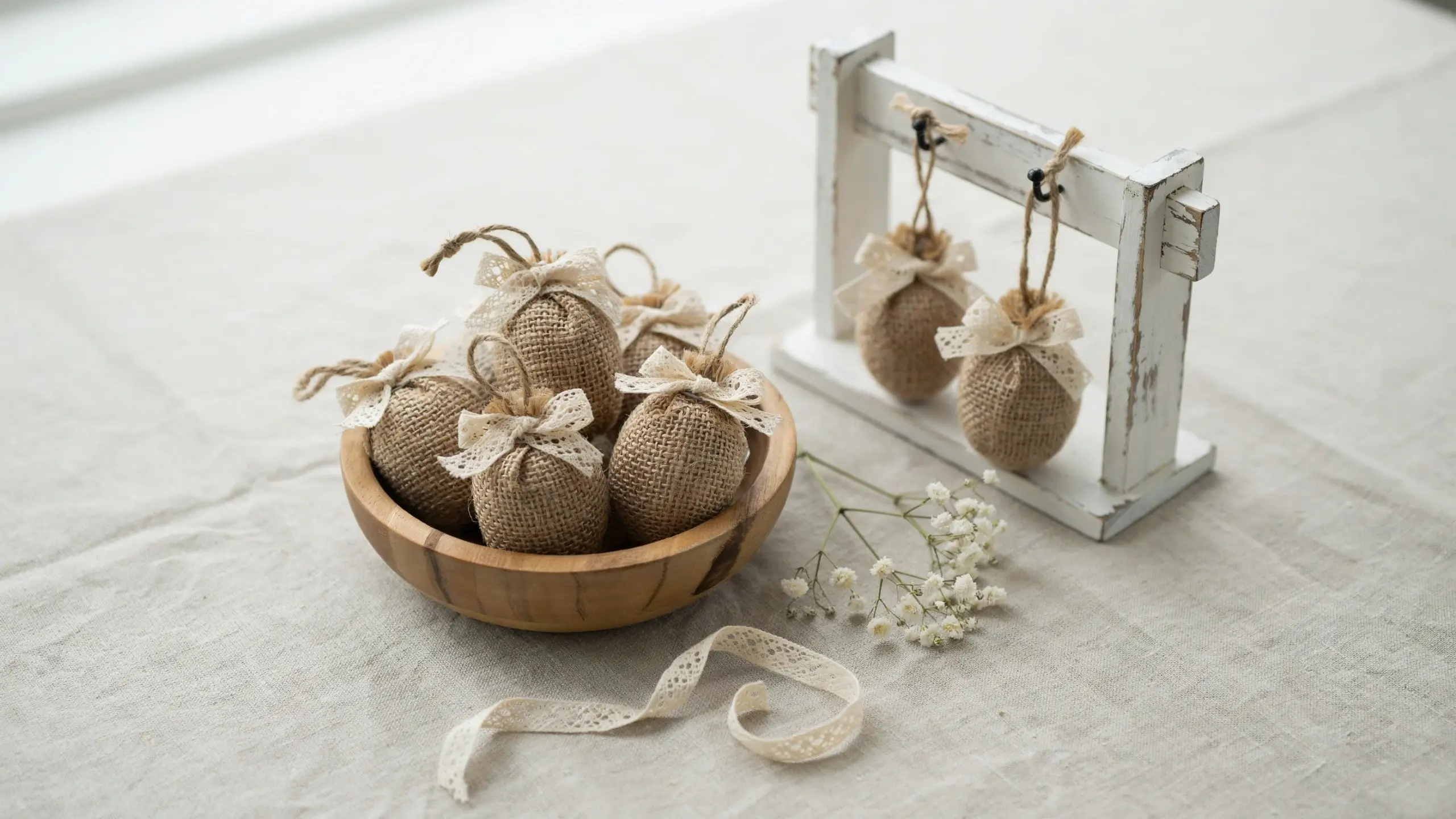 A collection of rustic burlap and lace-wrapped egg ornaments clustered in a light wooden bowl, with two pieces hanging from a distressed white painted display stand, resting on a beige linen tablecloth beside dried baby's breath and a lace ribbon.