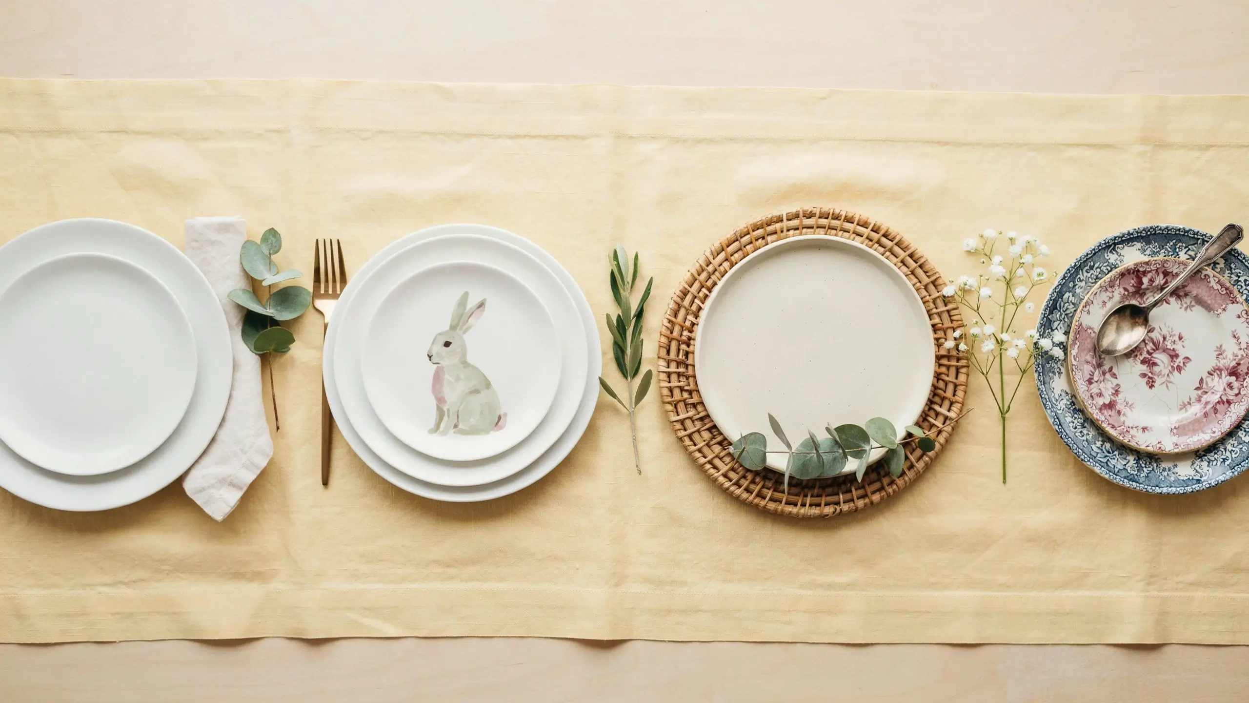 A curated rustic spring table setting featuring a mix of white ceramic plates, a bunny motif dish, a woven rattan charger, gold cutlery, and baby's breath on a yellow linen table runner.