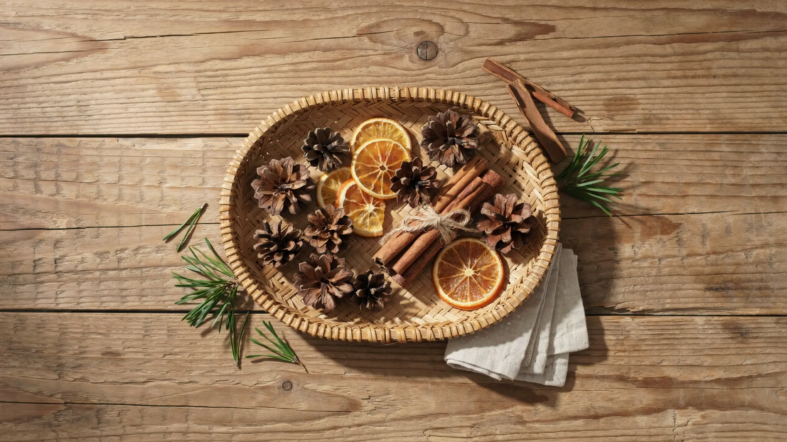 A woven round wicker tray filled with dried orange slices, pinecones, and cinnamon sticks tied with twine on a rustic wooden table, surrounded by pine needles and a folded white napkin.