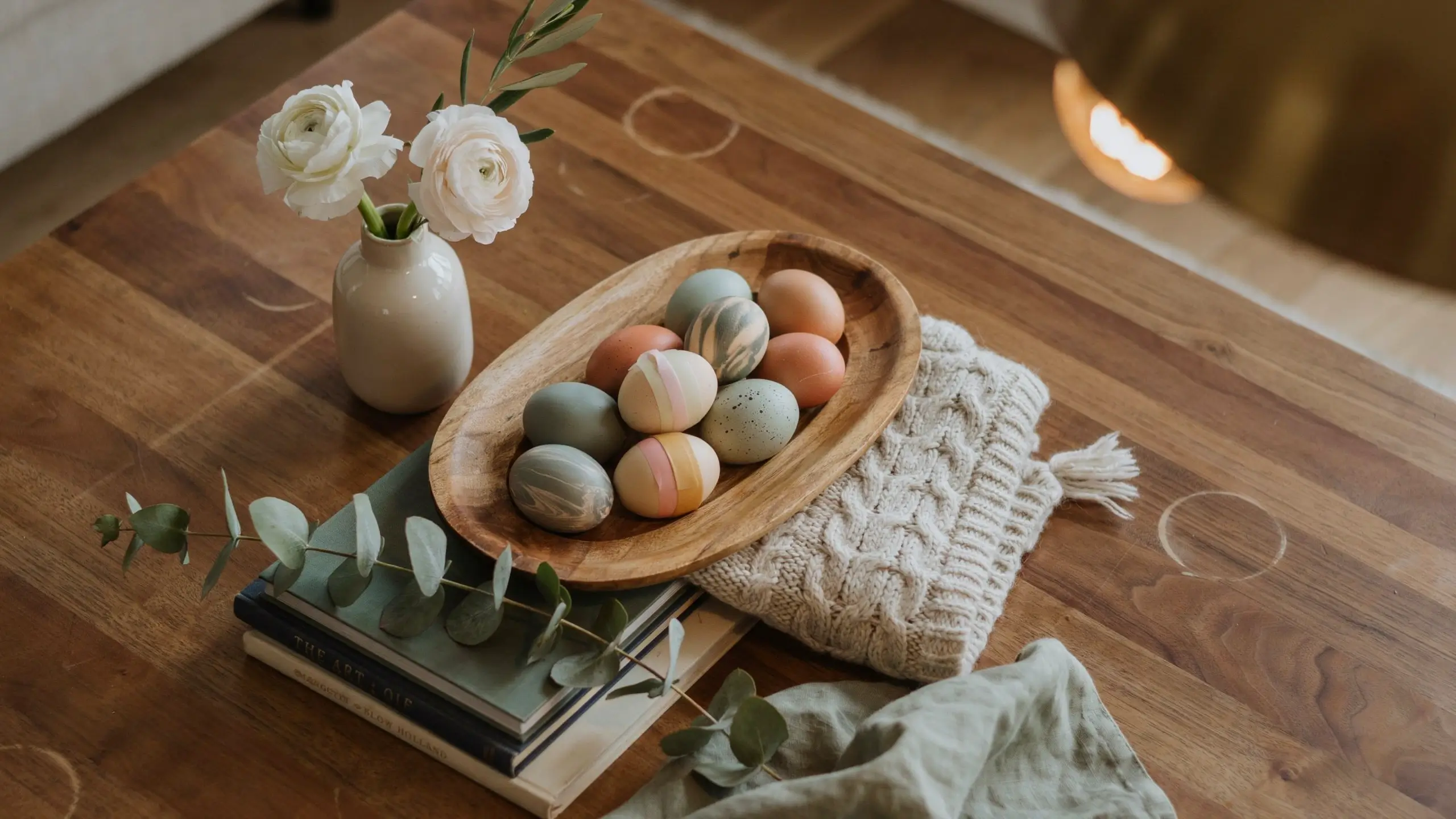 Rustic wooden oval tray filled with hand-dyed pastel eggs in sage green, peach, and cream, placed on a stack of books next to a white ceramic vase with white ranunculus flowers and a cream knit blanket.