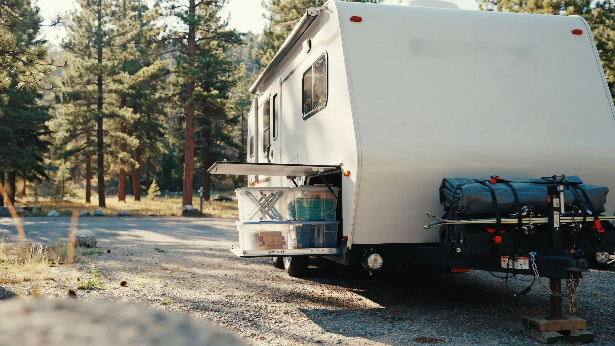 White RV in pine forest with open side storage bay of bins and rear hitch carrier hauling bulky tent gear.