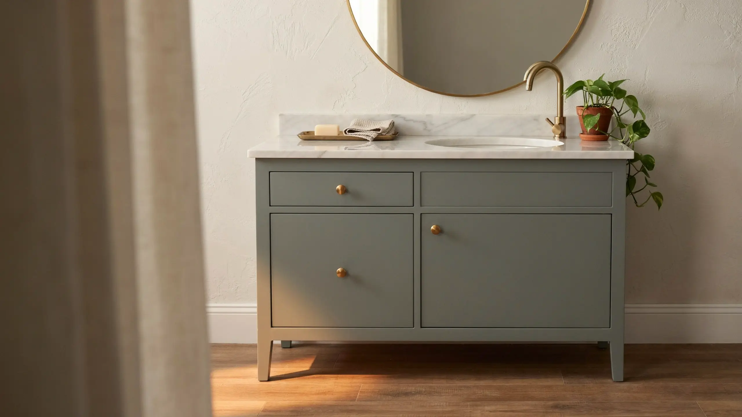 Modern sage green freestanding bathroom vanity featuring brass knobs, a round gold mirror, a white marble countertop, and a potted pothos plant illuminated by natural light.