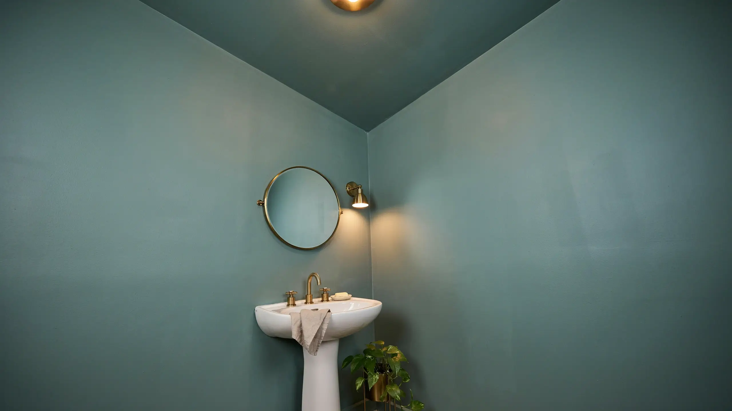 Bathroom corner featuring a white pedestal sink with a brass widespread faucet, round brass mirror, and wall sconce. The walls and ceiling are painted a cohesive, soothing sage green to create an immersive, spa-like atmosphere.