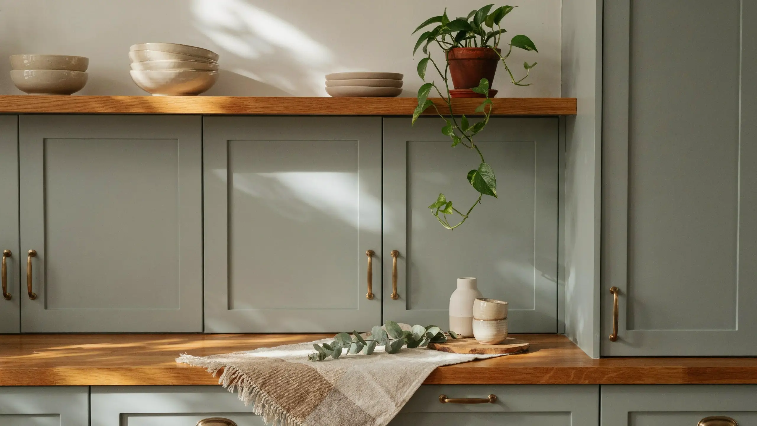 Sage green shaker kitchen cabinets with natural wood open shelving and a butcher block countertop, styled with a terracotta pothos plant, stacked ceramic bowls, a white vase, and a fringed linen runner bathed in warm diagonal sunlight.