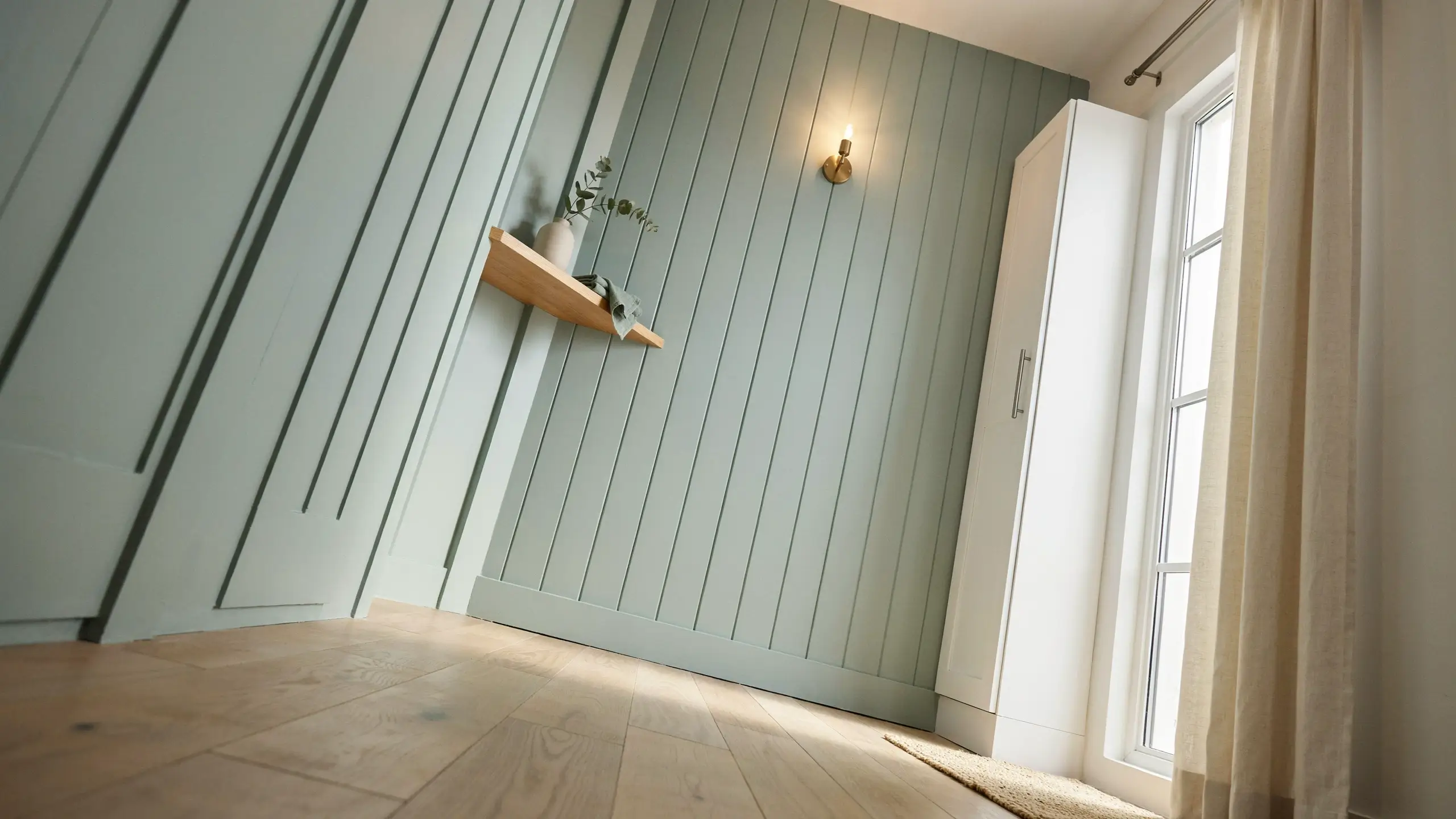 Sage green vertical shiplap paneling on a slanted attic wall with a wooden floating shelf, white vase, and warm wall sconce, demonstrating how vertical lines pull the eye upward to maximize ceiling height.