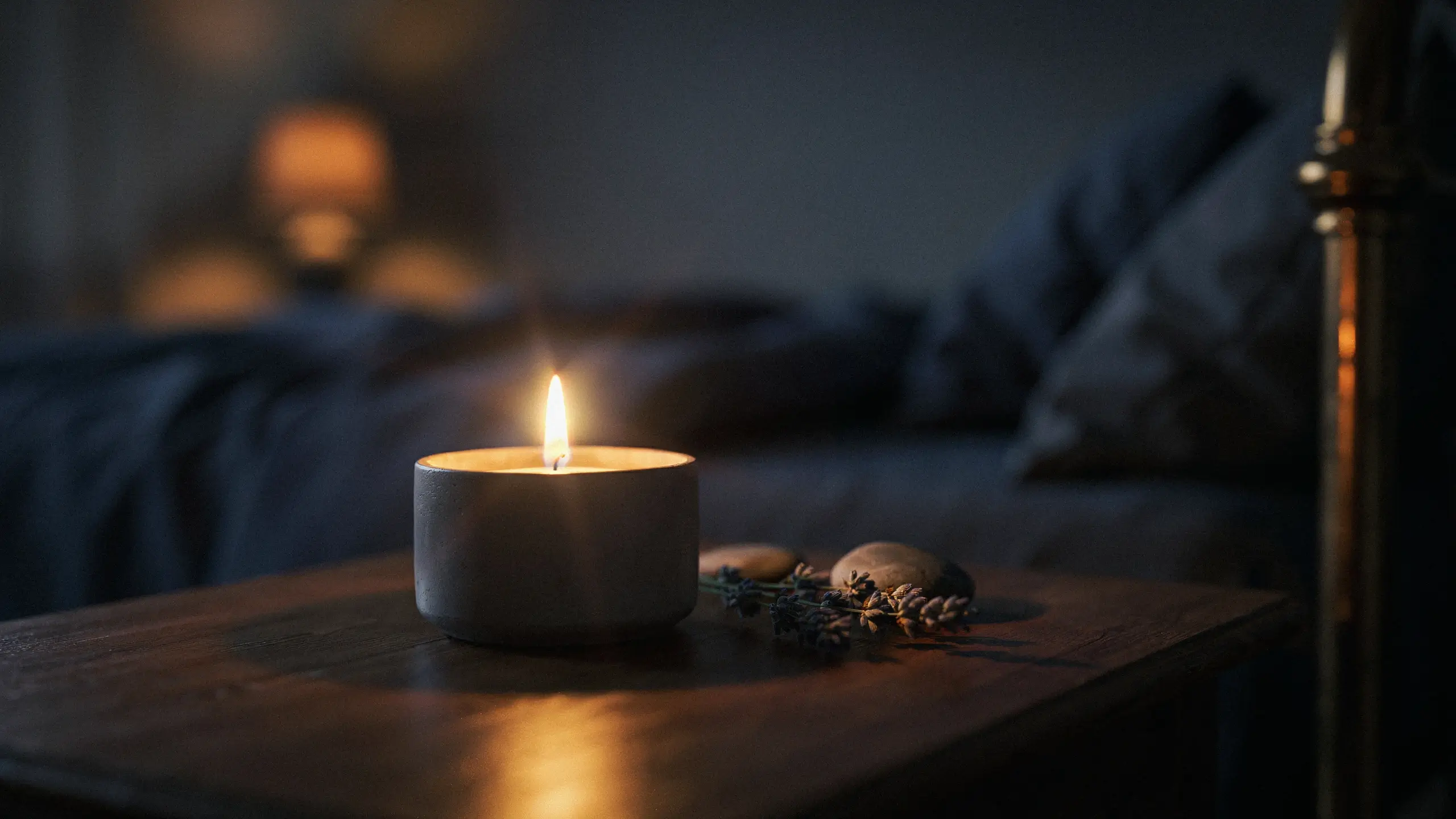 A lit white candle in a jar with dried lavender and stones set on a wooden nightstand to create a relaxing evening ambiance in a dark bedroom.