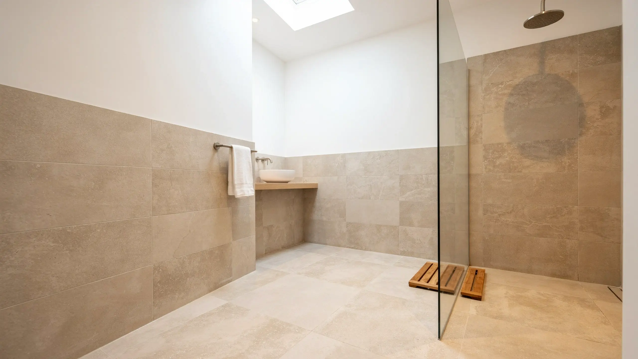 Modern bathroom showcasing seamless beige stone floor-to-wall tiles, featuring a floating wooden vanity with a white vessel sink and a glass shower partition demonstrating continuous tile design.