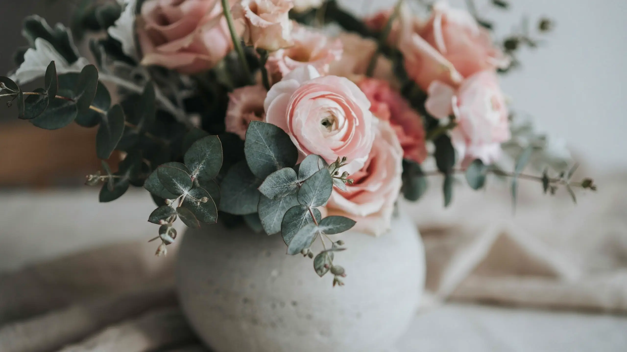 Pale blush ranunculus and seeded eucalyptus bouquet in a round white textured ceramic vase on a draped linen surface.