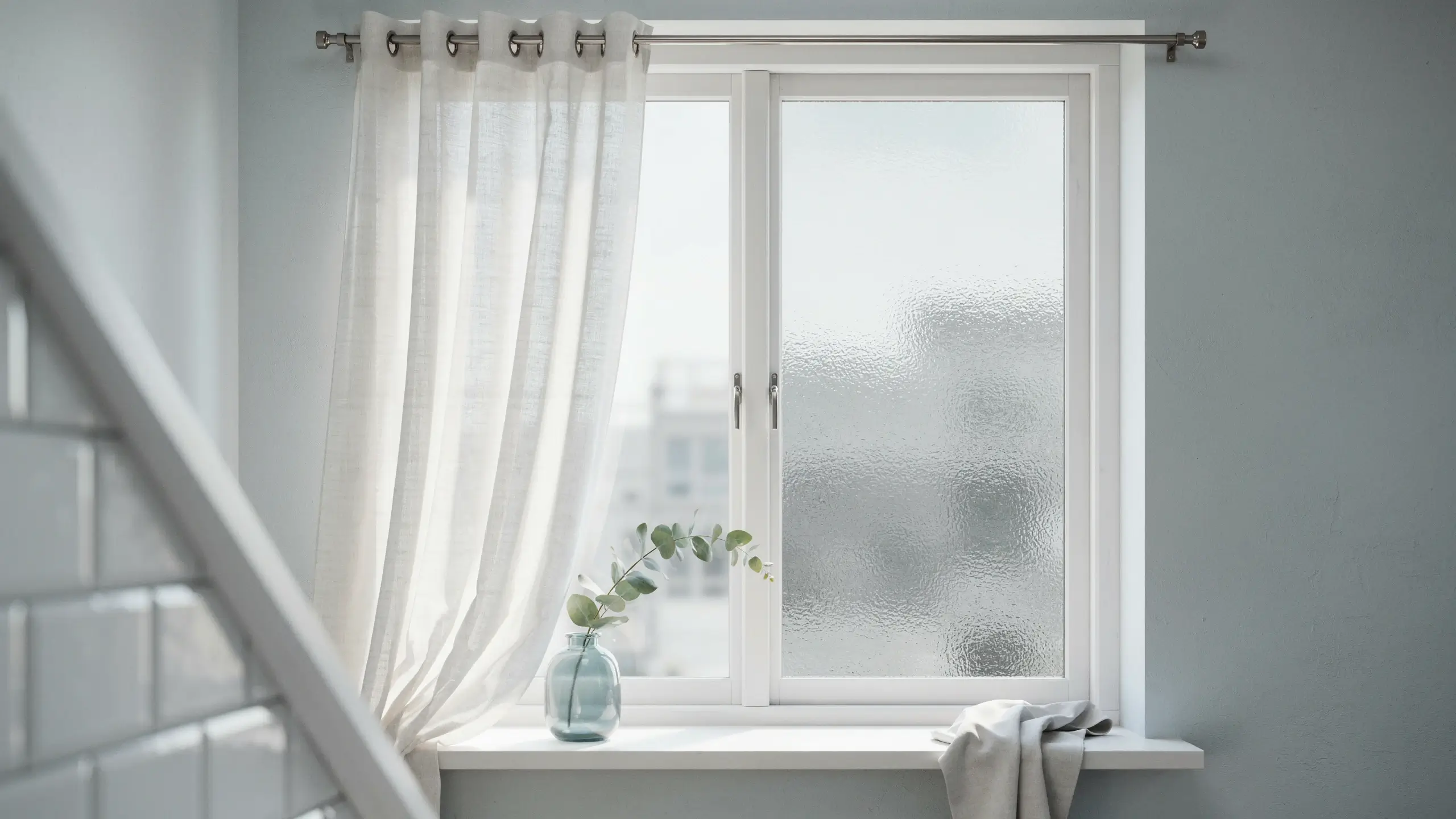 A bright minimalist window featuring sheer white curtains and a frosted glass pane, highlighting an alternative window treatment for natural light and privacy, with a blue glass vase and eucalyptus sprig on the white windowsill.