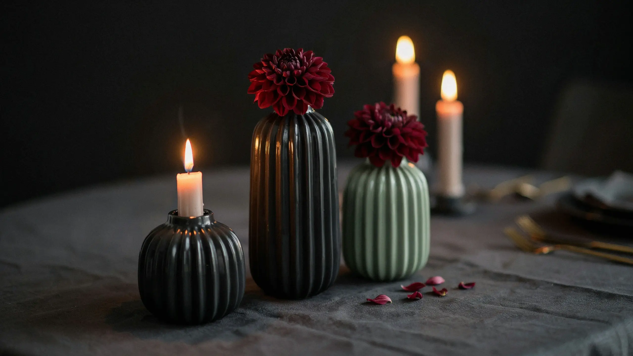 Two ribbed SKURAR ceramic vases spray painted in black and sage green holding deep red dahlias, paired with a matching black vase supporting a lit white taper candle, arranged on a dark gray linen tablecloth with scattered petals for a romantic moody tablescape.