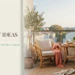 A cozy small balcony decorated with a rattan armchair, white cushions, and a pink throw blanket. An olive tree and snake plant bring greenery to the space, next to a small table holding a mug and an open book. The scene is lit by string lights with text reading "25 Small Balcony Ideas