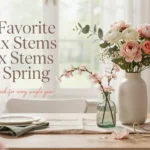 White ceramic vase holding faux pink peonies and cherry blossom stems on a bright wooden dining table with elegant spring place settings.