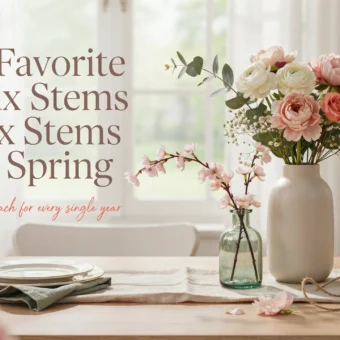 White ceramic vase holding faux pink peonies and cherry blossom stems on a bright wooden dining table with elegant spring place settings.