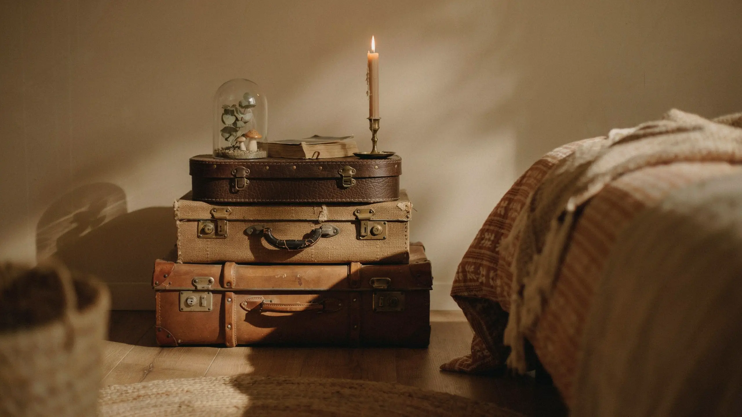 Three stacked vintage leather and canvas suitcases used as a rustic bedside table. On top sits a glass cloche holding dried foliage, a lit taper candle in a brass holder, and a closed book, set in a cozy, warmly lit bedroom. 