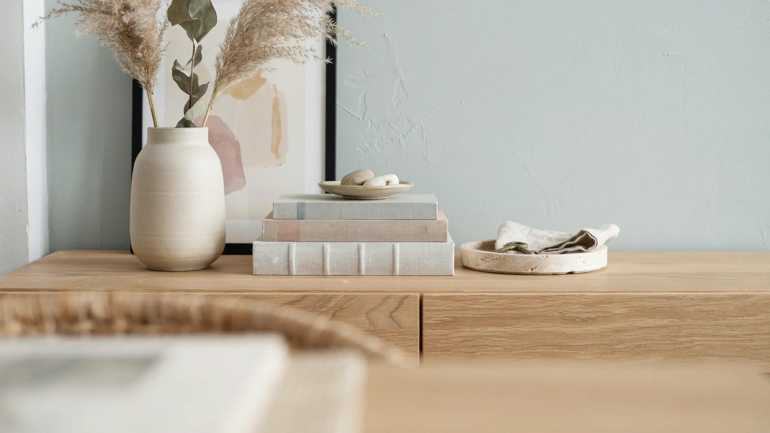 A neutral Japandi-style console table styling featuring a cream ceramic vase with dried pampas grass, a stack of linen-bound books with a small decorative dish, and a travertine tray with a cloth napkin.