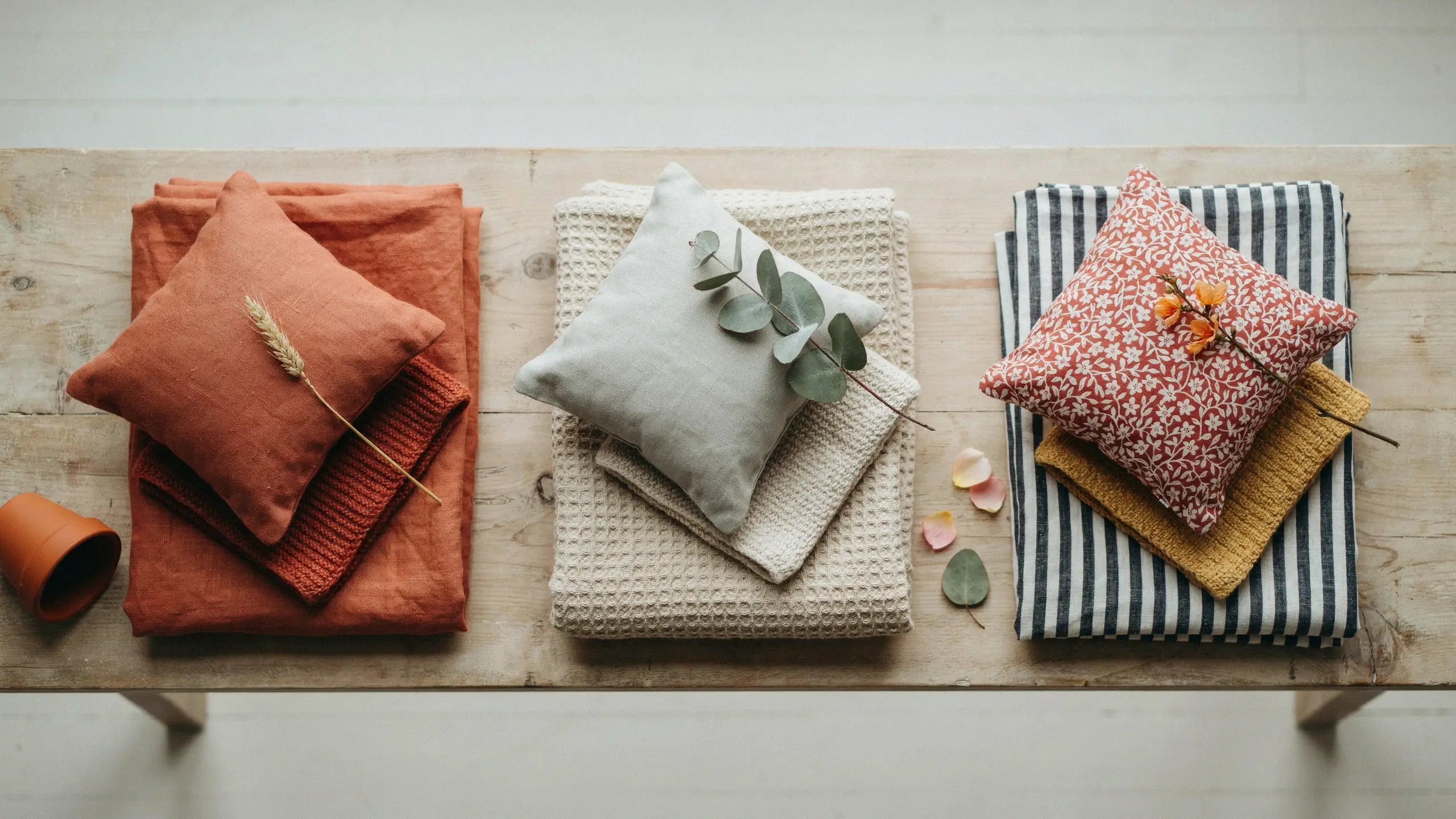 Three seasonal textile arrangements on a rustic wooden bench featuring rust orange fall pillows with dried wheat, neutral beige winter throws with eucalyptus, and spring floral cushions atop mustard and navy striped fabrics, accented by a small terracotta pot and scattered petals.