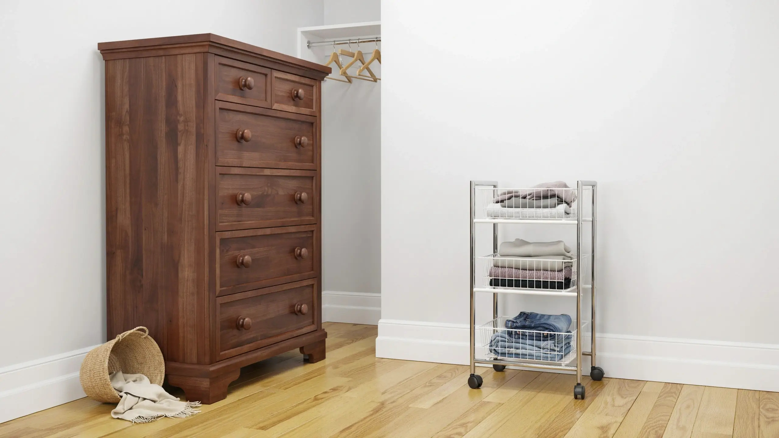 Tall dark wood chest of drawers next to a white wire rolling cart holding folded jeans and sweaters in a bright bedroom closet.
