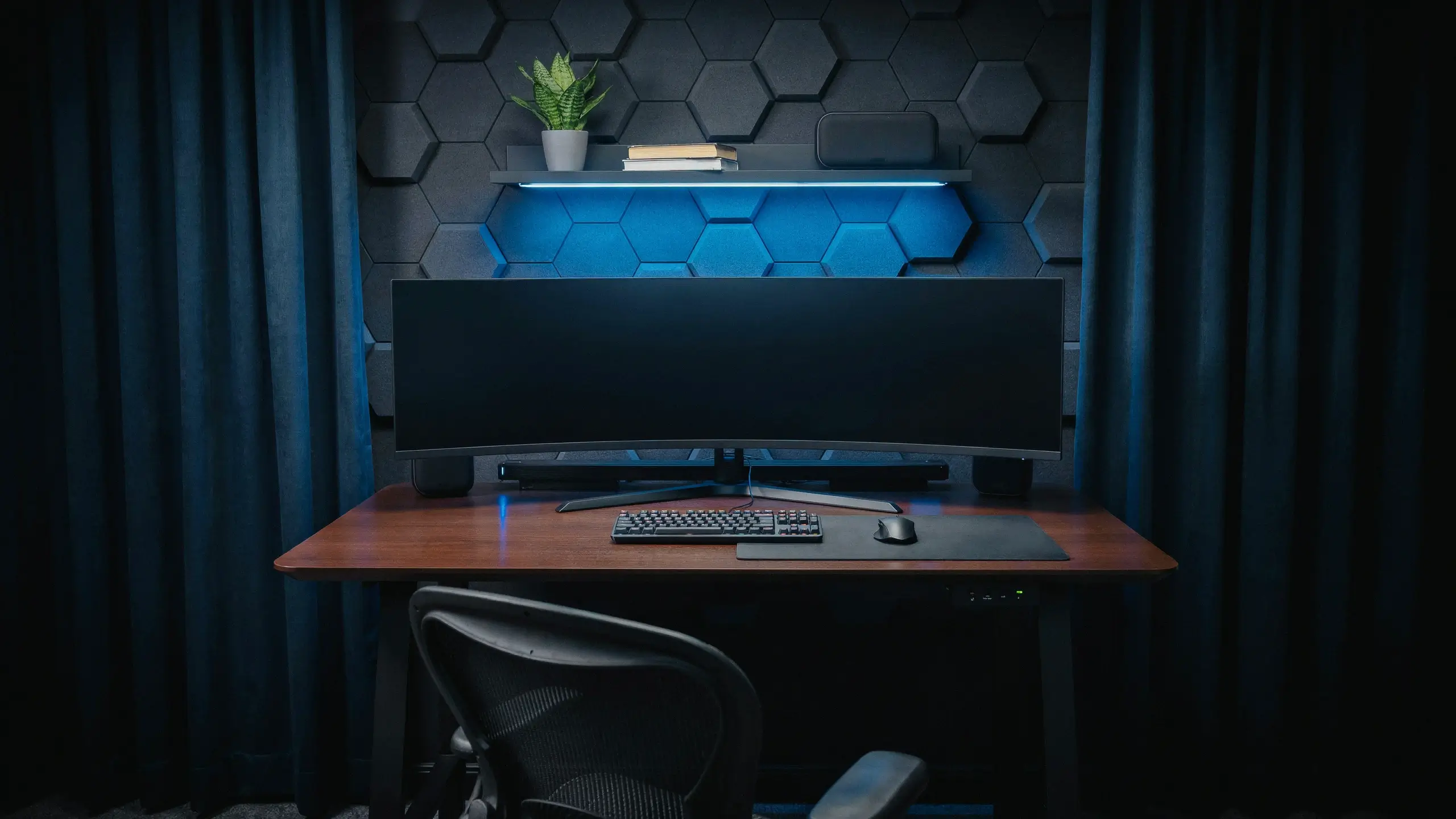 Modern tech-forward gamer den featuring a curved ultrawide monitor, ergonomic mesh chair, and blue LED-lit hexagonal acoustic wall panels.
