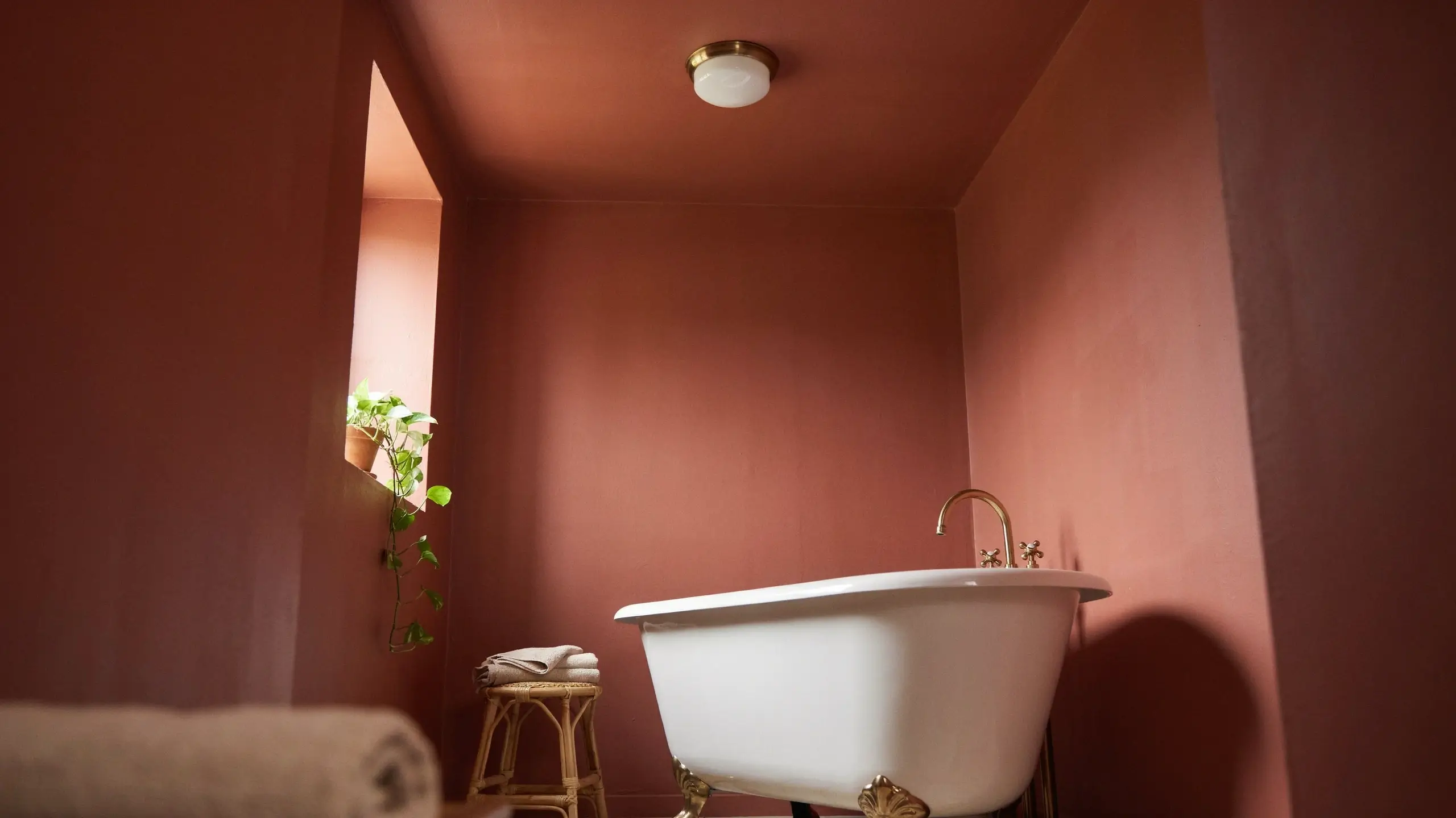 A cozy terracotta bathroom featuring a painted ceiling that matches the walls, a white clawfoot tub with elegant brass hardware, a rattan stool with stacked towels, and a trailing pothos plant in a recessed window niche.