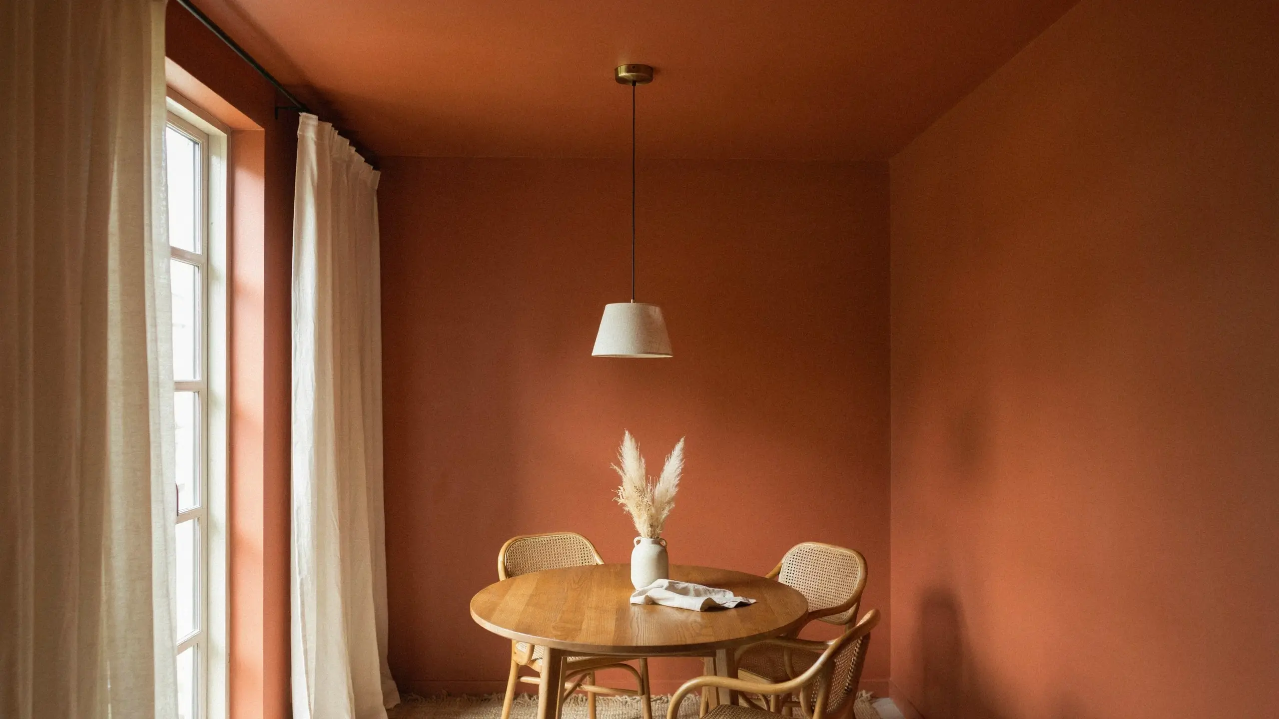 Warm monochromatic terracotta dining room with a painted ceiling, featuring a round oak table, cane-back chairs, white pendant lamp, and a vase of pampas grass.