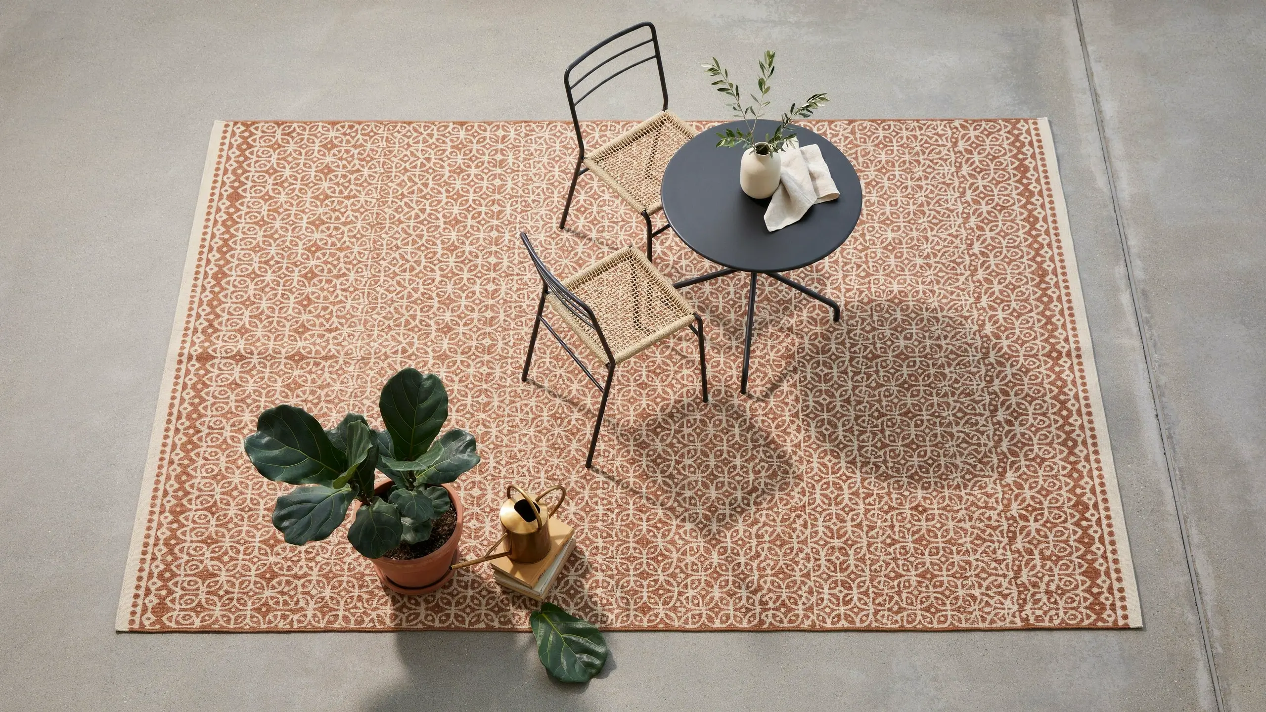 A terracotta patterned outdoor rug anchoring a modern patio setup with black metal chairs, woven cane seats, a round table, and a potted fiddle leaf fig plant.