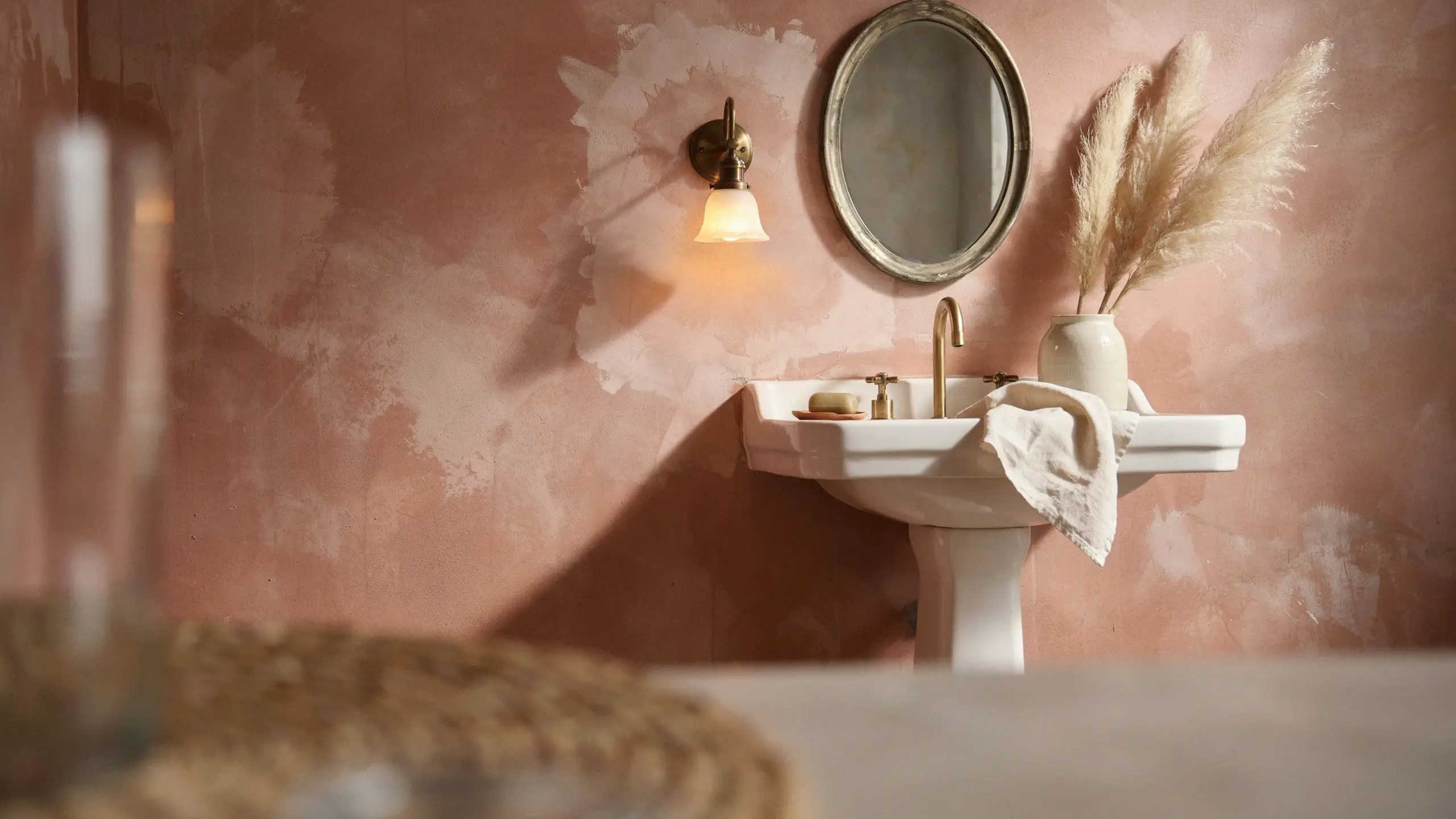 A close-up of a warm, peach-toned limewash or Venetian plaster textured wall in a bathroom, featuring a white pedestal sink with a brass faucet, a round mirror, a brass wall sconce, and a vase of dried pampas grass.