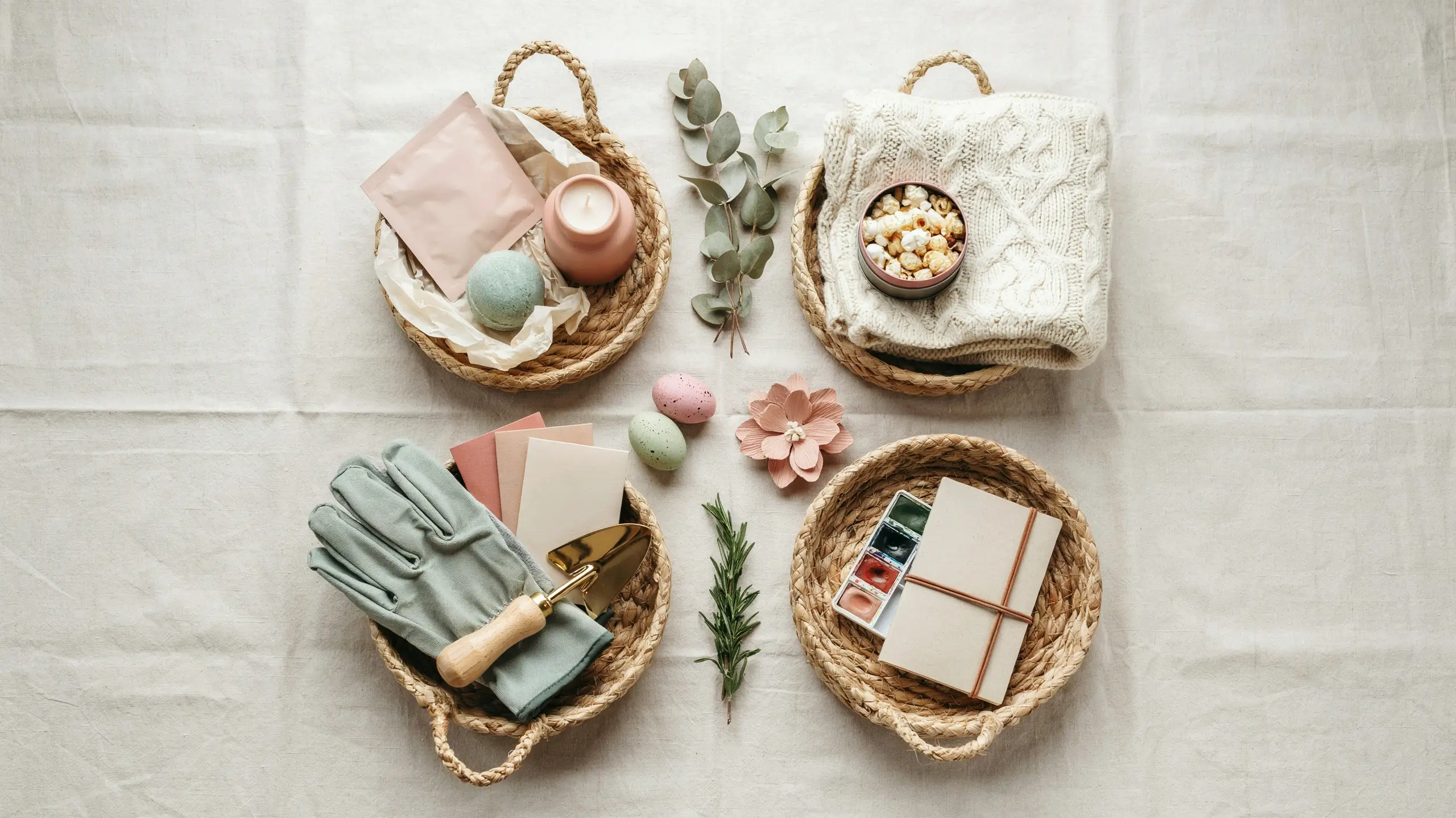 Flat lay of four thoughtfully themed Easter baskets with useful gifts including self-care items, a cozy snack tin, gardening tools, and watercolor art supplies on a white linen background.