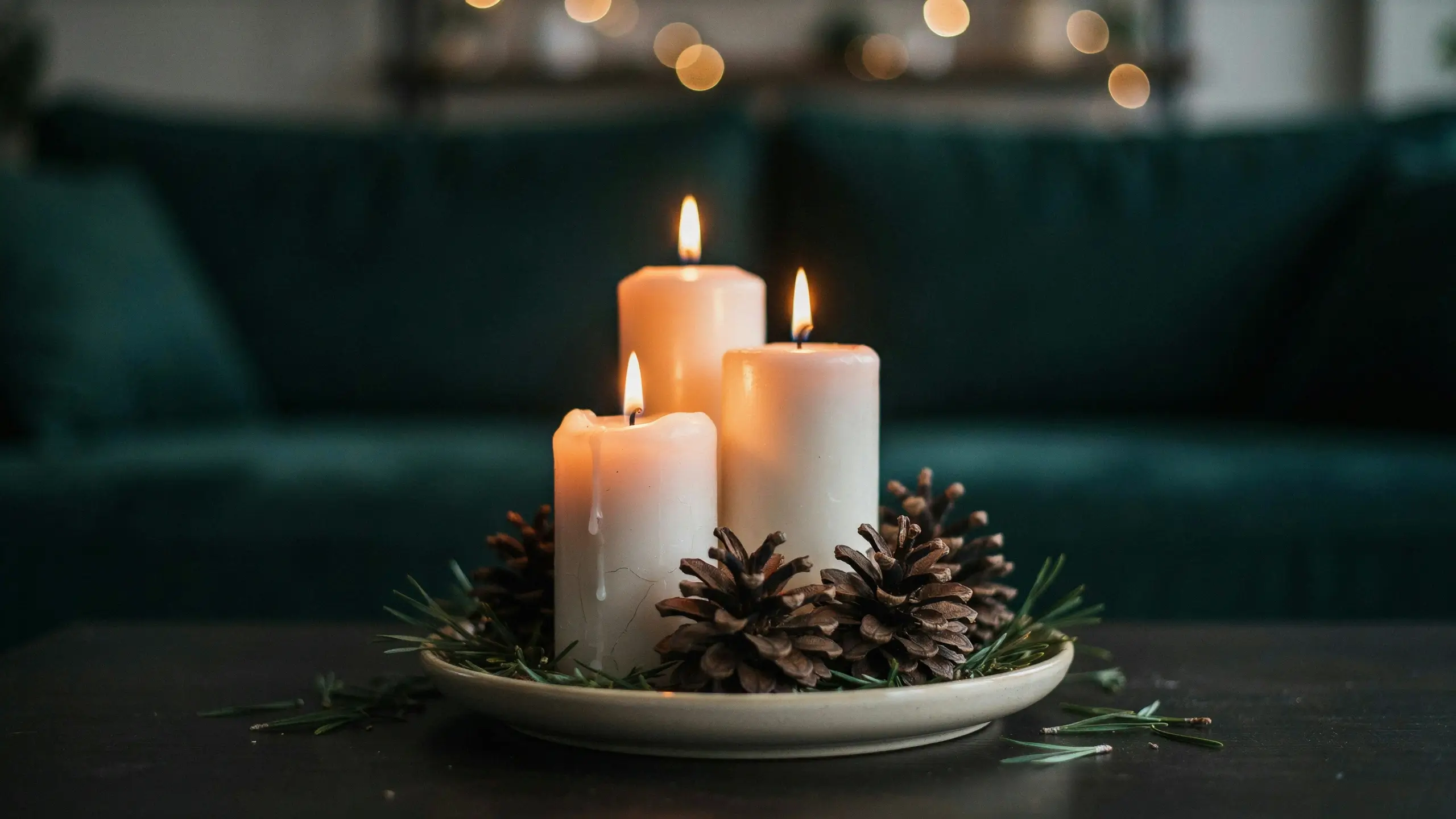 Three lit white pillar candles with pine cones and evergreen sprigs on a white plate, creating a cozy winter centerpiece.