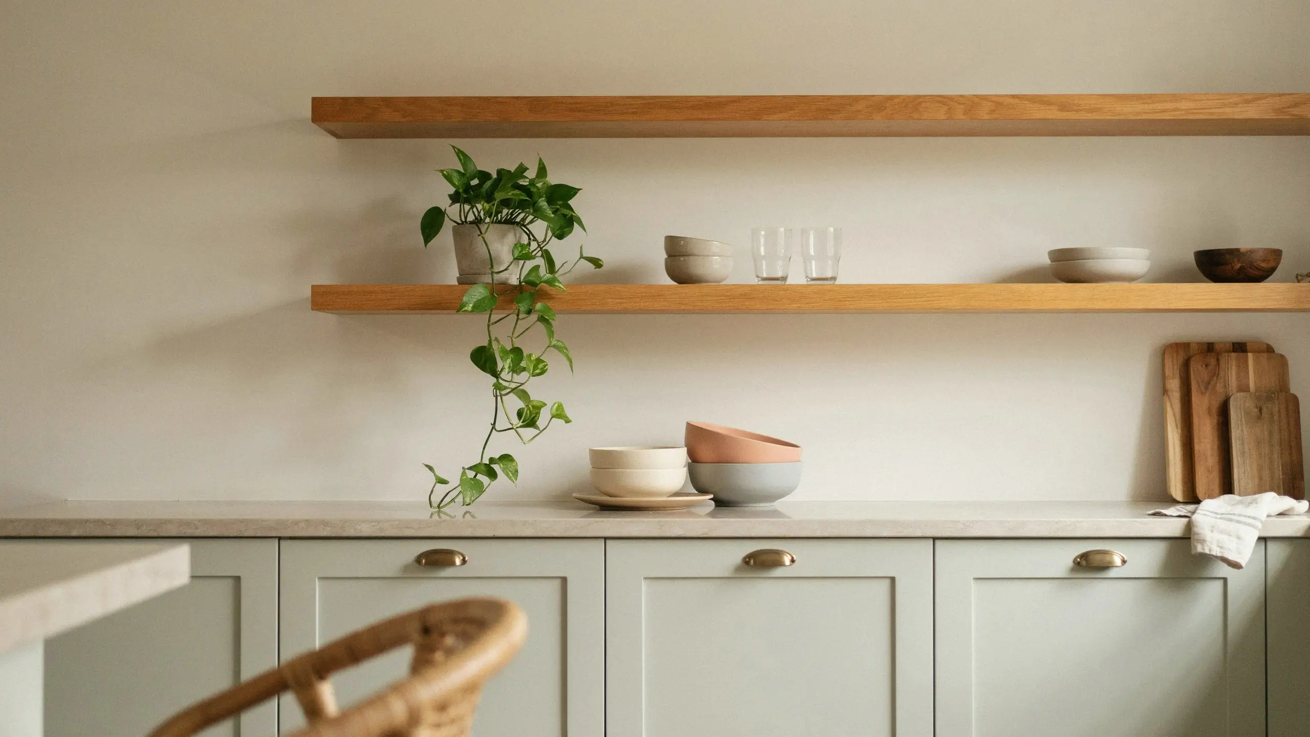 Sage green classic Shaker-style kitchen cabinets with brushed brass cup pulls, paired with warm wood floating shelves holding a trailing potted pothos plant, stacked ceramic bowls, glass tumblers, and wooden cutting boards for a timeless minimalist design.