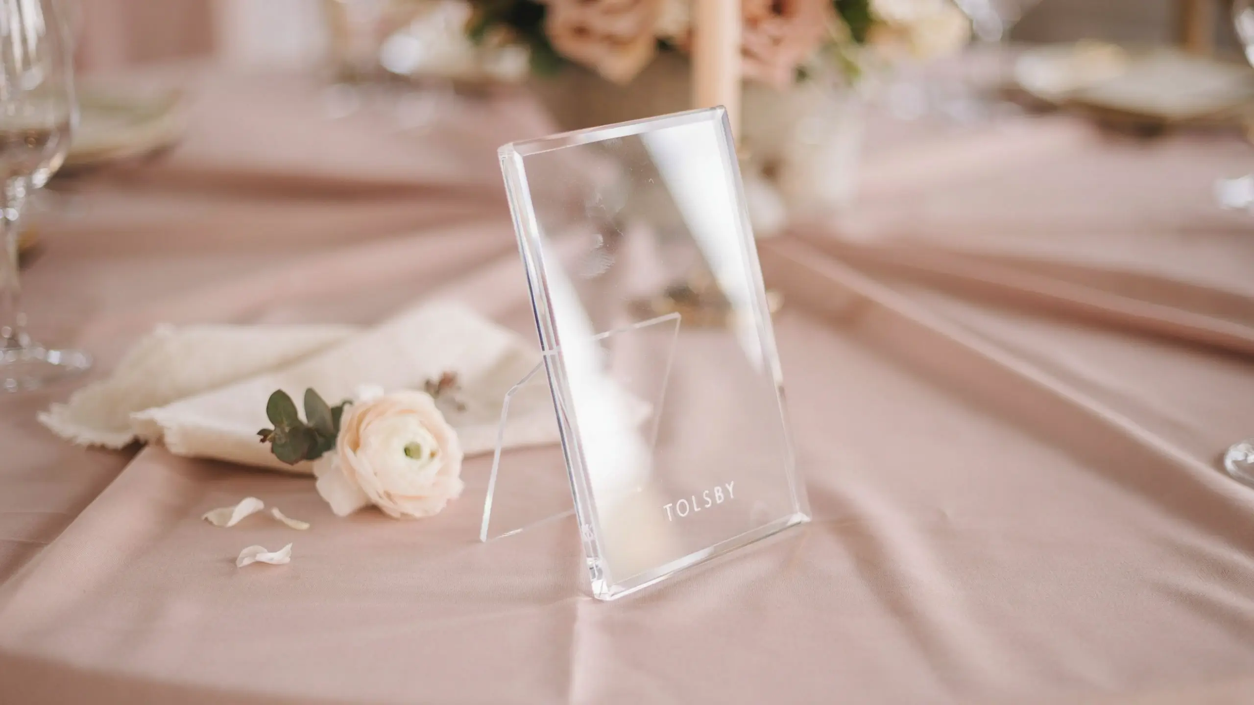 Clear TOLSBY acrylic table number frame standing upright on a dusty blush pink wedding tablecloth, placed next to a folded cream linen napkin and a soft pale ranunculus flower with scattered petals.