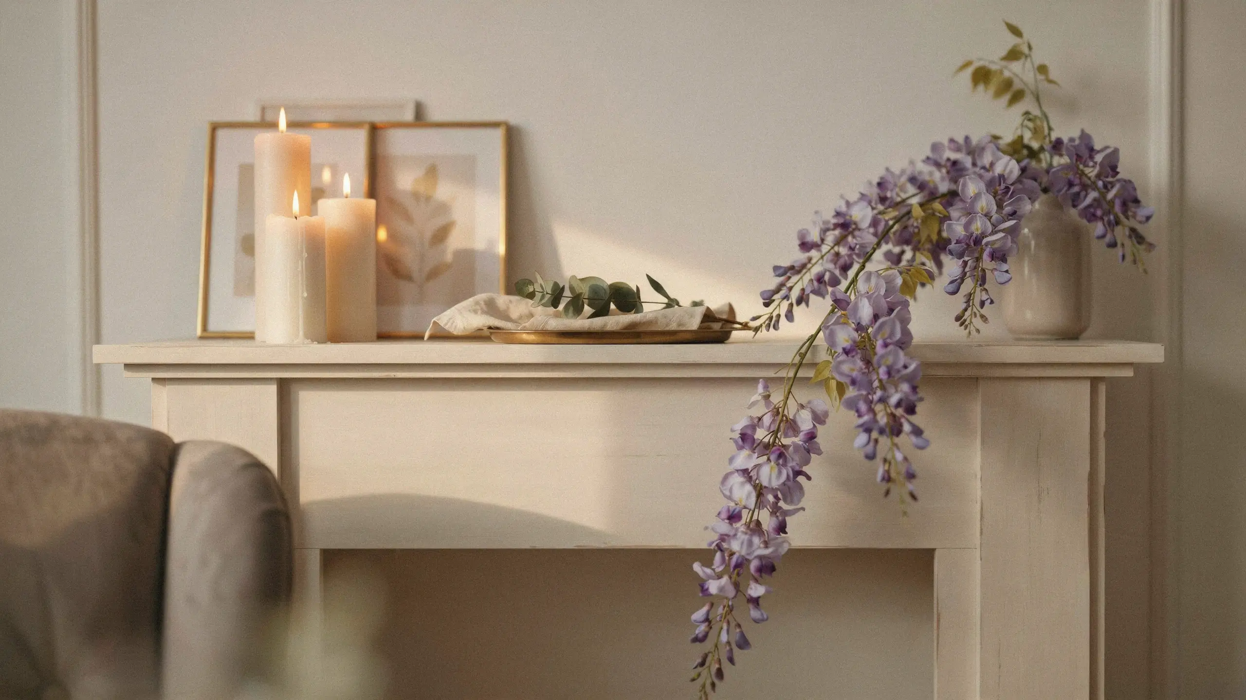 Trailing purple wisteria stems cascading gracefully from a matte beige vase on a white wooden console table, flanked by lit cream pillar candles and framed botanical prints, showcasing minimal spring home styling.