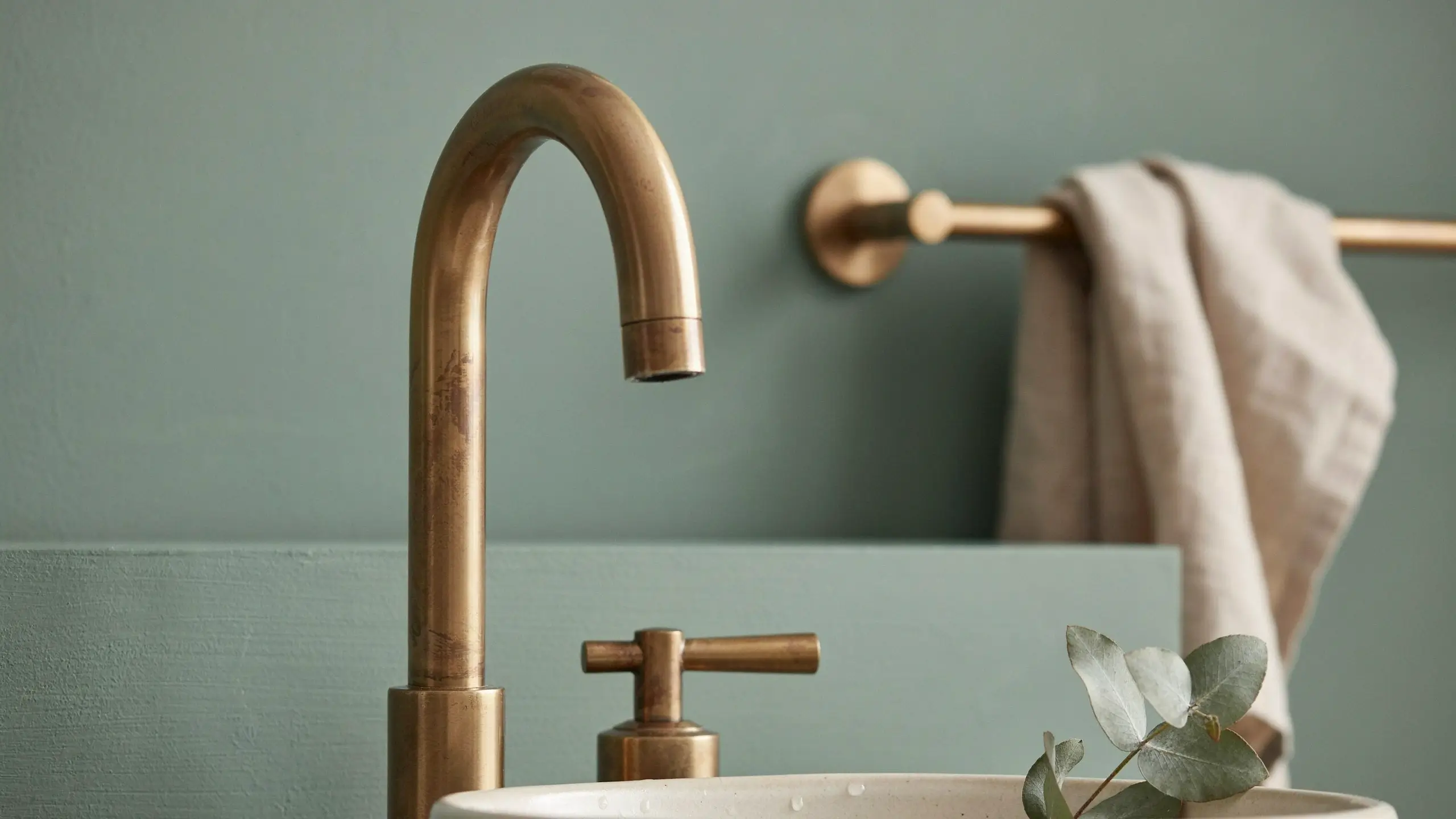A close-up of an unlacquered brass gooseneck faucet with a cross-handle mounted on a white basin, set against a soft sage green bathroom wall with a beige linen towel and a eucalyptus sprig.