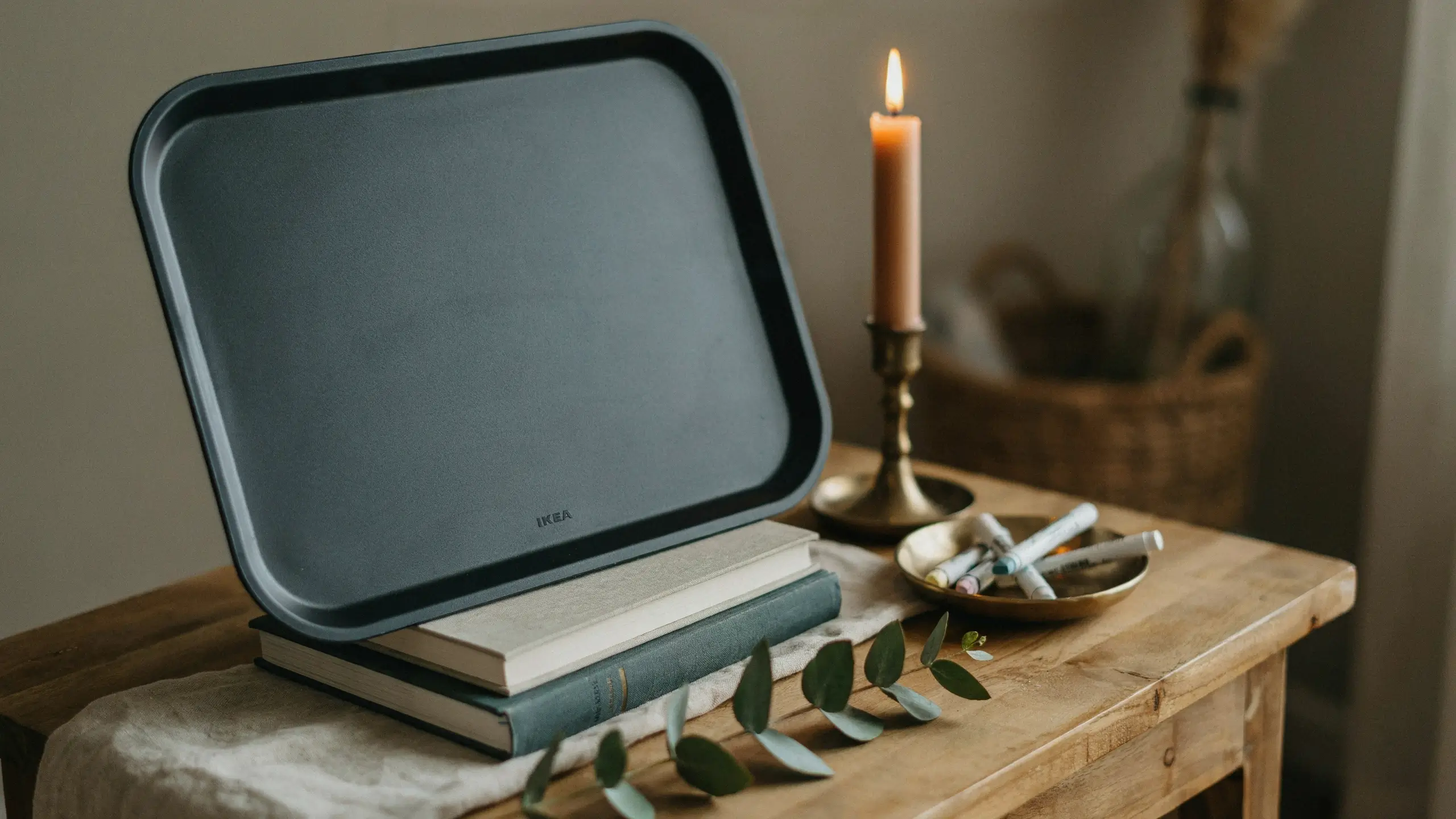 Dark grey IKEA tray propped on stacked books styled as a rustic DIY chalkboard sign, surrounded by a lit candle, a brass bowl of chalk pieces, and fresh eucalyptus leaves on a weathered wood table for cozy home decor.