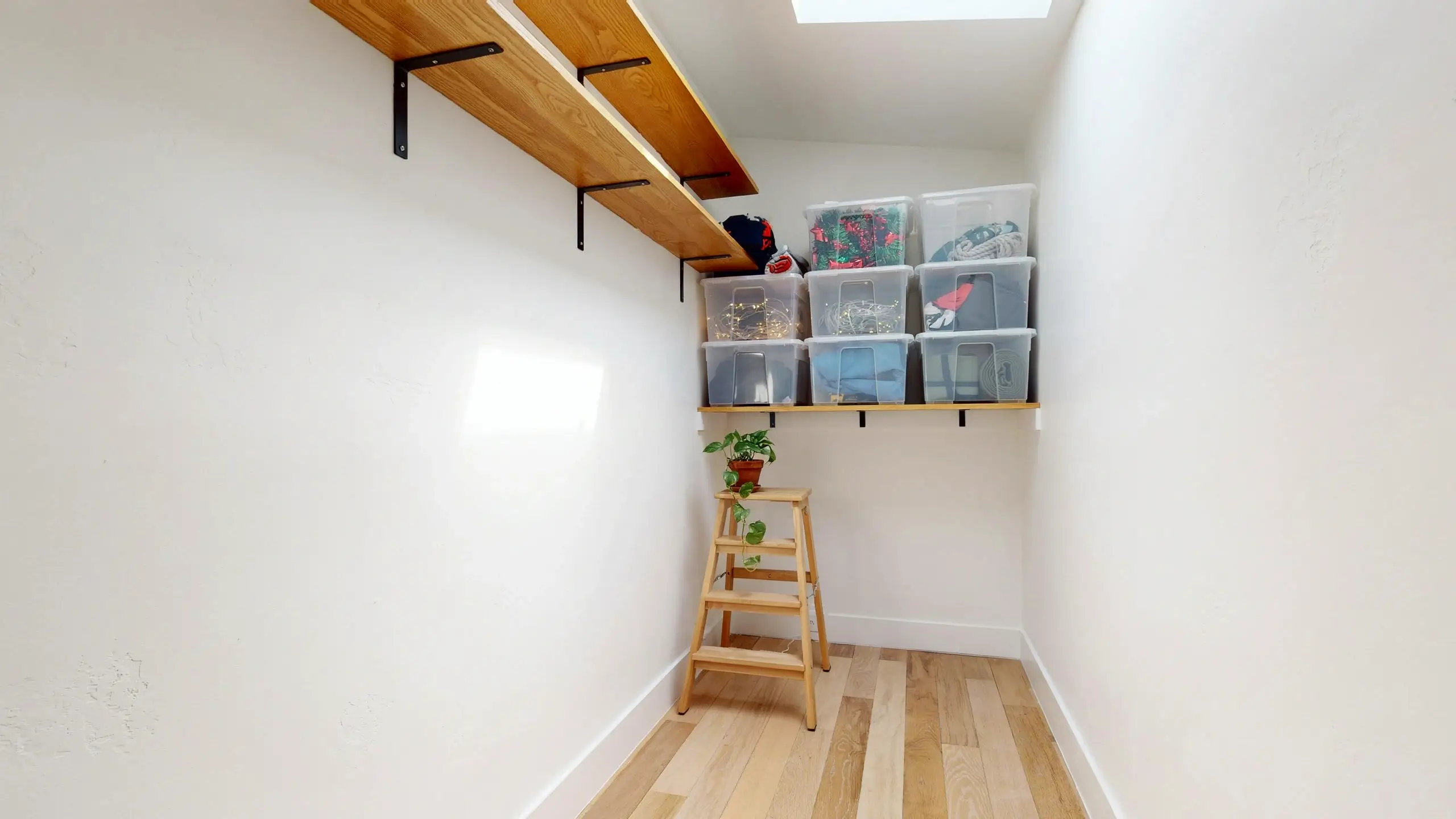 A white storage closet featuring high wooden shelves holding neat rows of clear plastic bins, with a wooden step stool placed in the center holding a small potted plant to easily access rarely-used seasonal items.