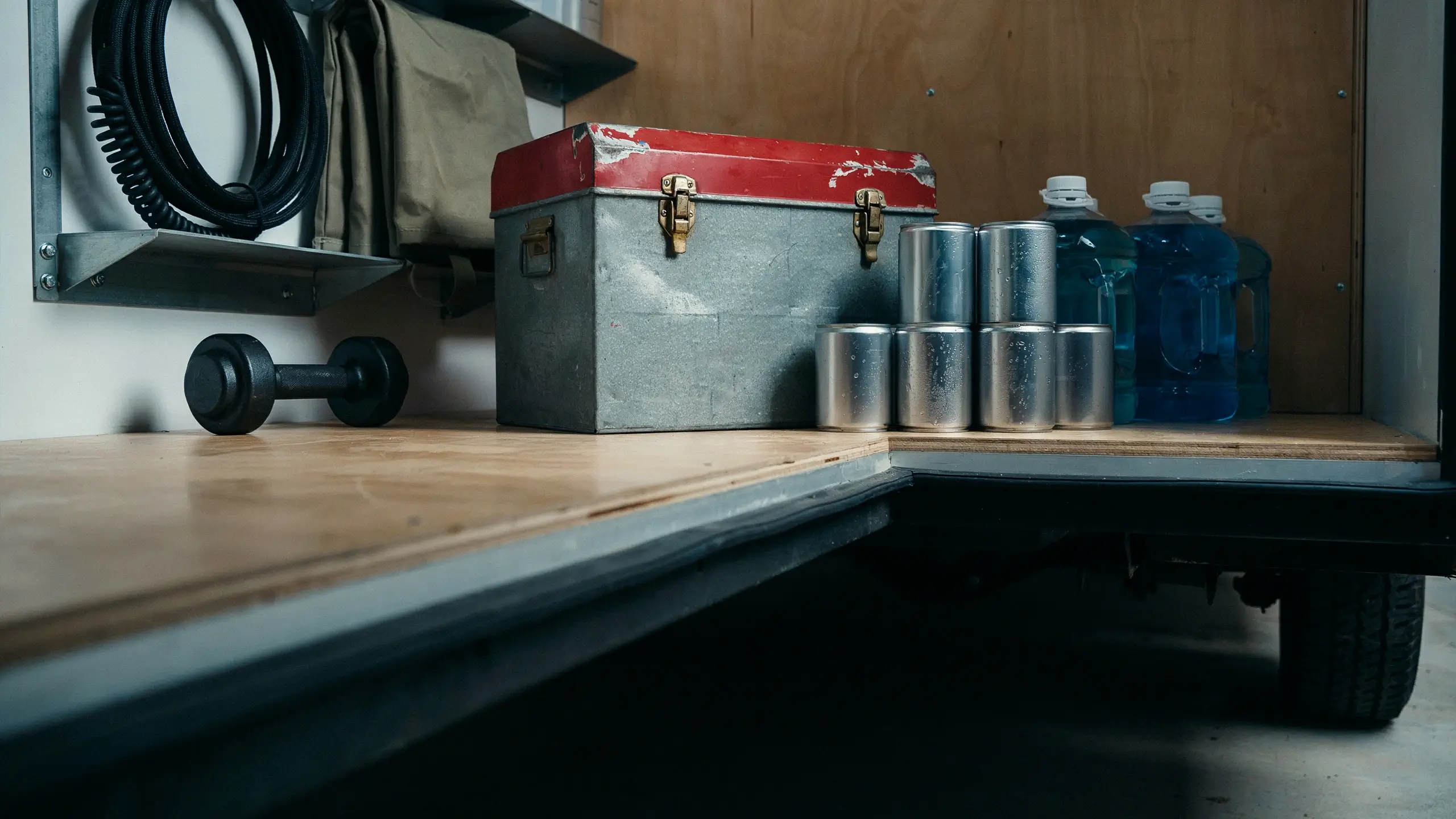 Heavy metal storage chest, blue water jugs, and fuel canisters loaded onto a wooden van floor to demonstrate balanced cargo weight distribution for safe overlanding handling.