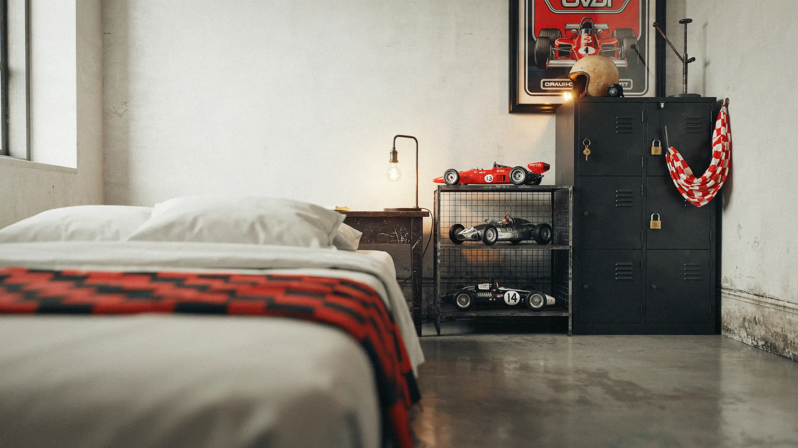 Vintage motorsport garage style bedroom with black metal lockers, a yellow racing helmet, a draped red-and-white checkered flag, and three retro model race cars displayed on a wire shelf.