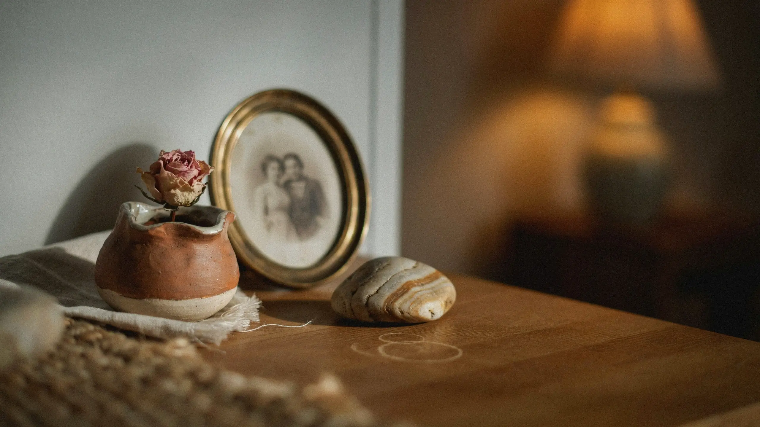 Vintage brass framed couple portrait, dried pink rose in a ceramic pot, and a striped stone on a wooden table with warm lighting. 