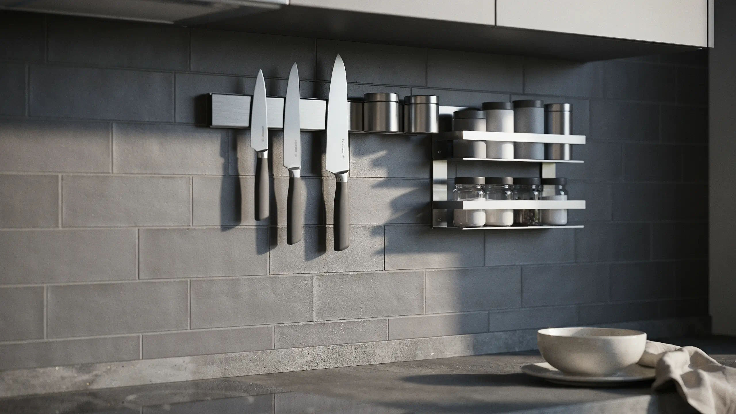 Three kitchen knives hanging on a sleek magnetic knife strip next to a two-tier stainless steel spice rack on a gray subway tile wall.