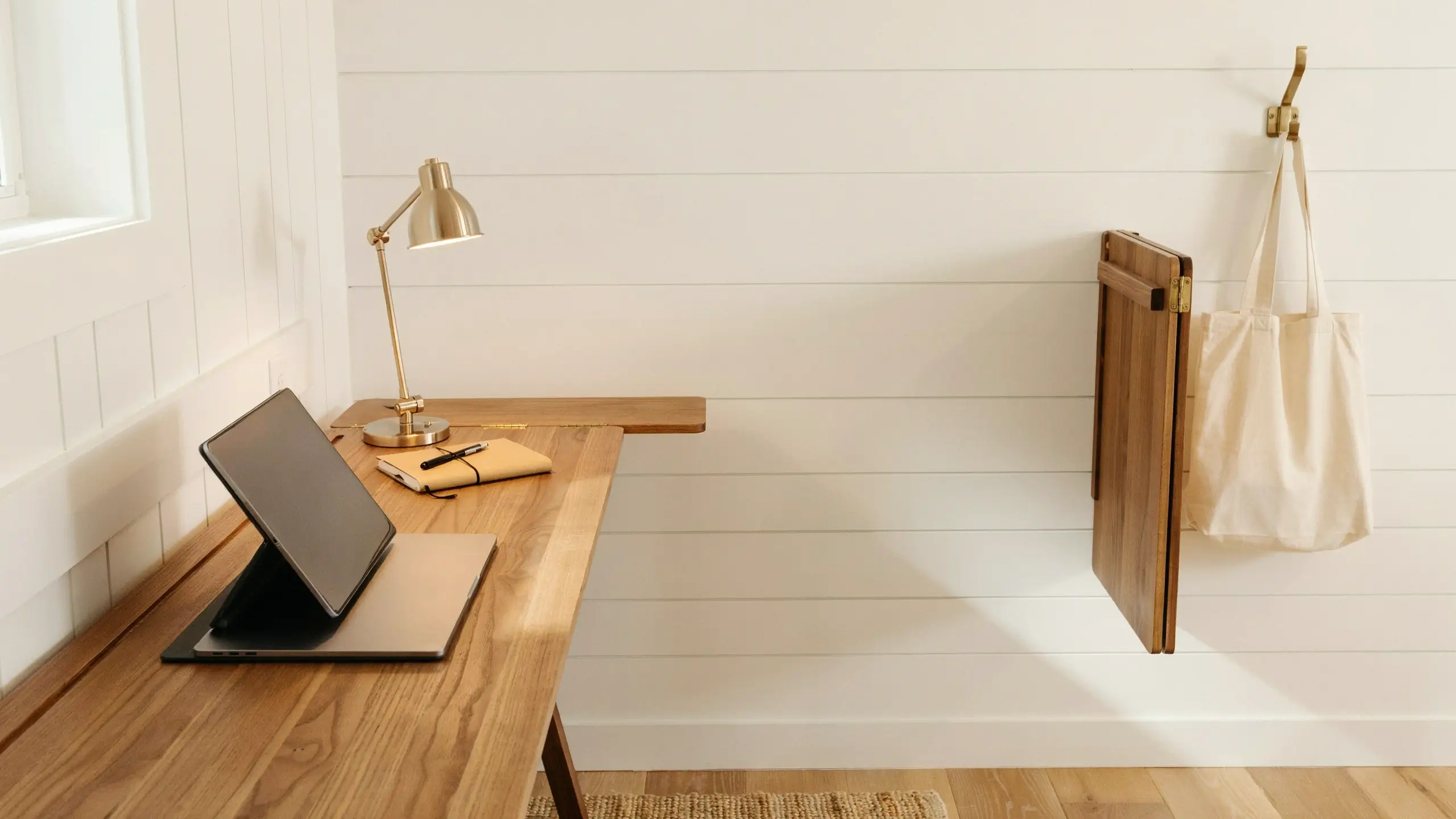 Space-saving wall-mounted wooden drop-leaf desk holding a brass lamp, notebook, and open tablet, designed for a minimalist home office corner.