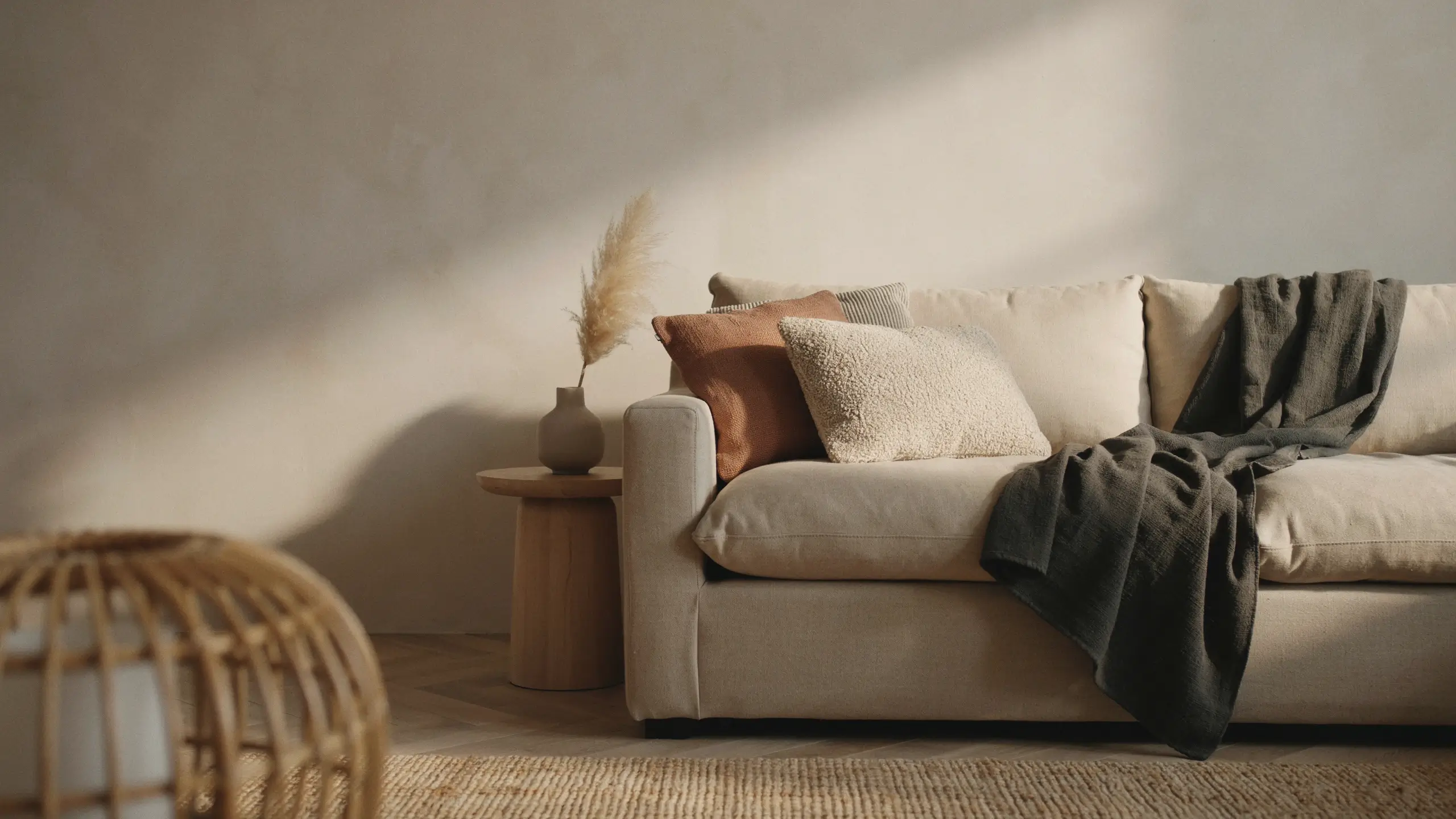 Warm neutral minimalist living room corner featuring a plush cream beige sofa styled with a terracotta bouclé pillow and a dark grey textured throw blanket, paired with dried pampas grass in a ceramic vase on a wooden side table, bathed in soft natural sunlight.