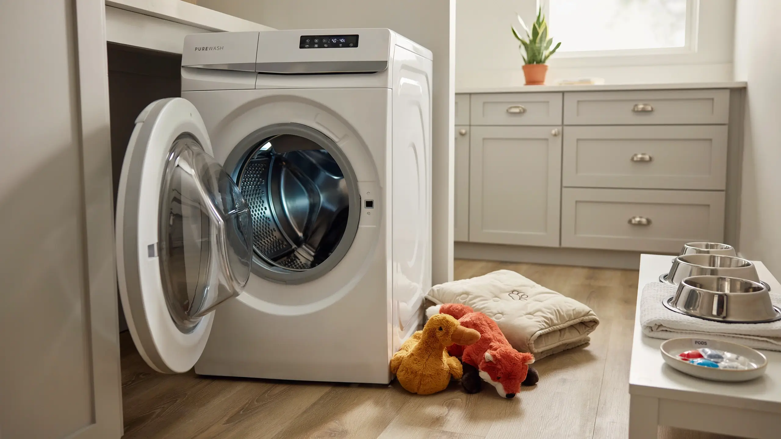 Open white washing machine in a bright laundry room, ready to sanitize pet bedding, a stuffed duck and fox toy, and metal dog bowls, highlighting a regular cleaning schedule for pet accessories.