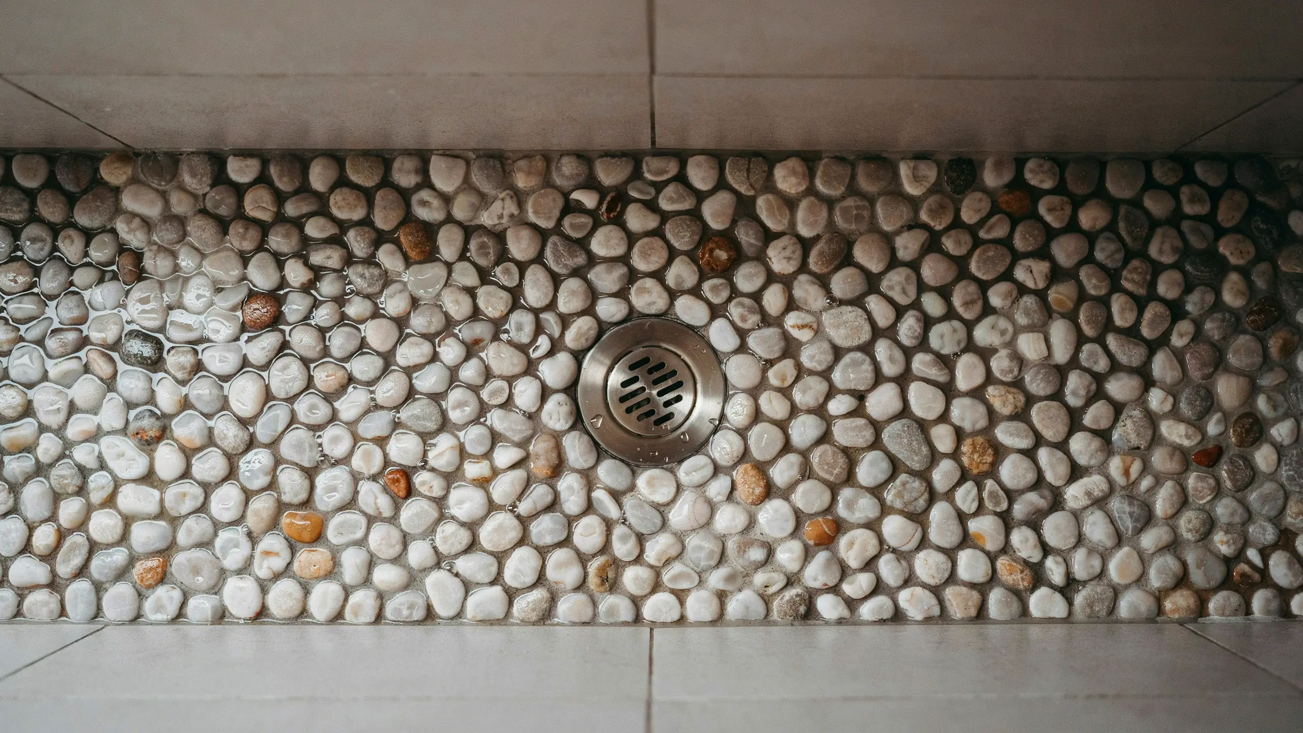 Close-up view of a wet pebble stone shower floor featuring a central stainless steel floor drain, surrounded by large white ceramic tiles.