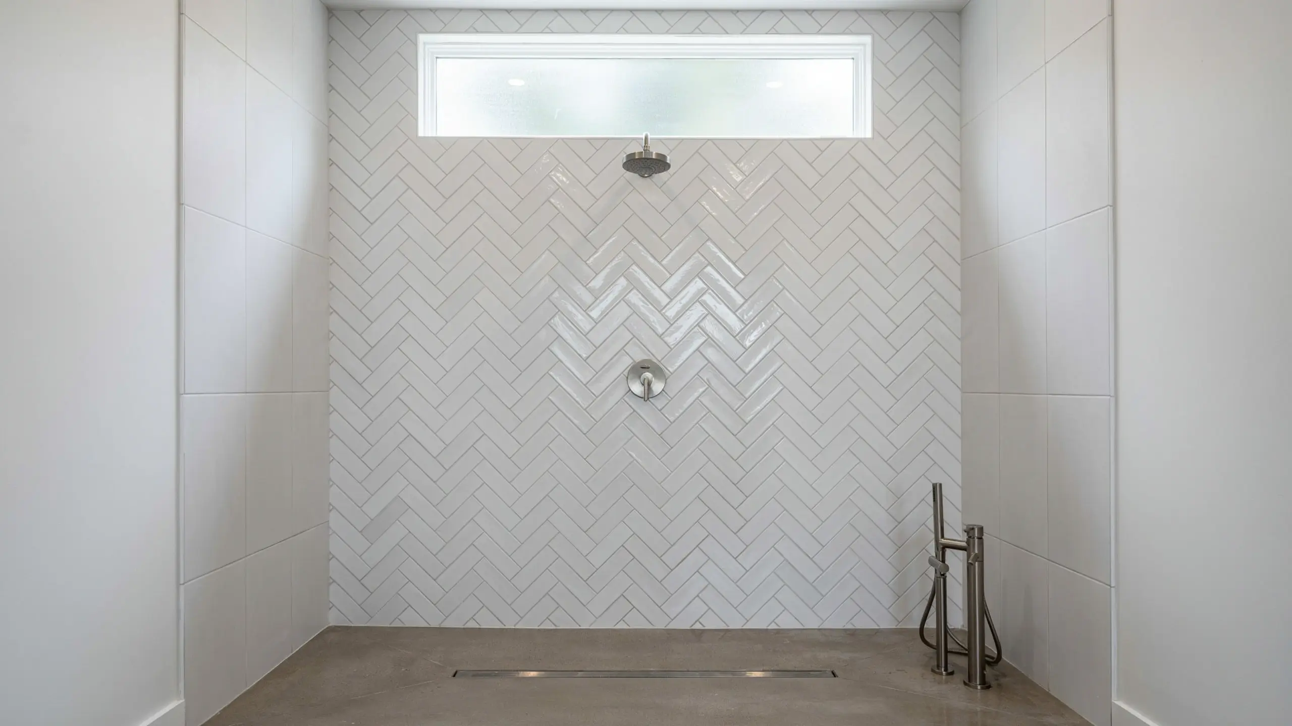 Modern walk-in shower featuring a white herringbone tile accent wall behind the brushed nickel showerhead and control valve, complete with a linear floor drain and a high transom window.