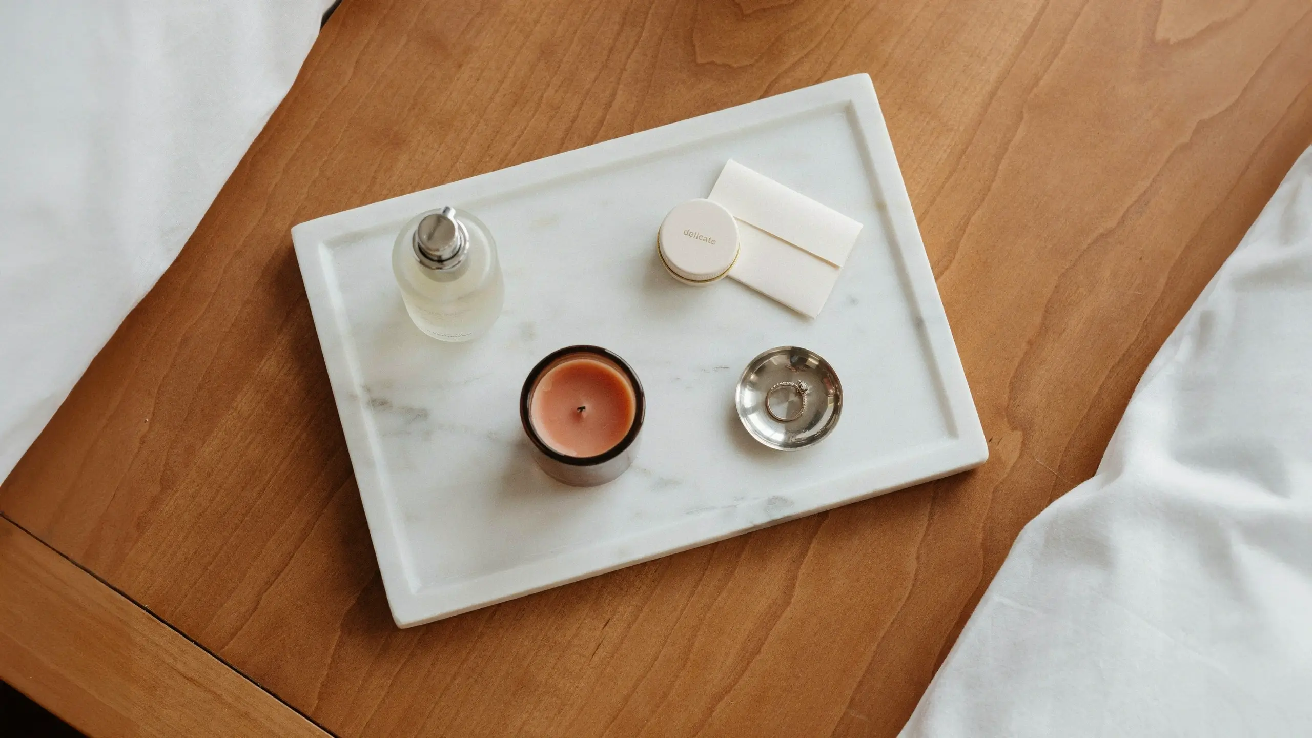 A minimalist white marble tray organizing a glass perfume bottle, a 'delicate' cream jar, a folded napkin, a candle in a black metal case, and a silver dish on a warm wooden hotel nightstand.