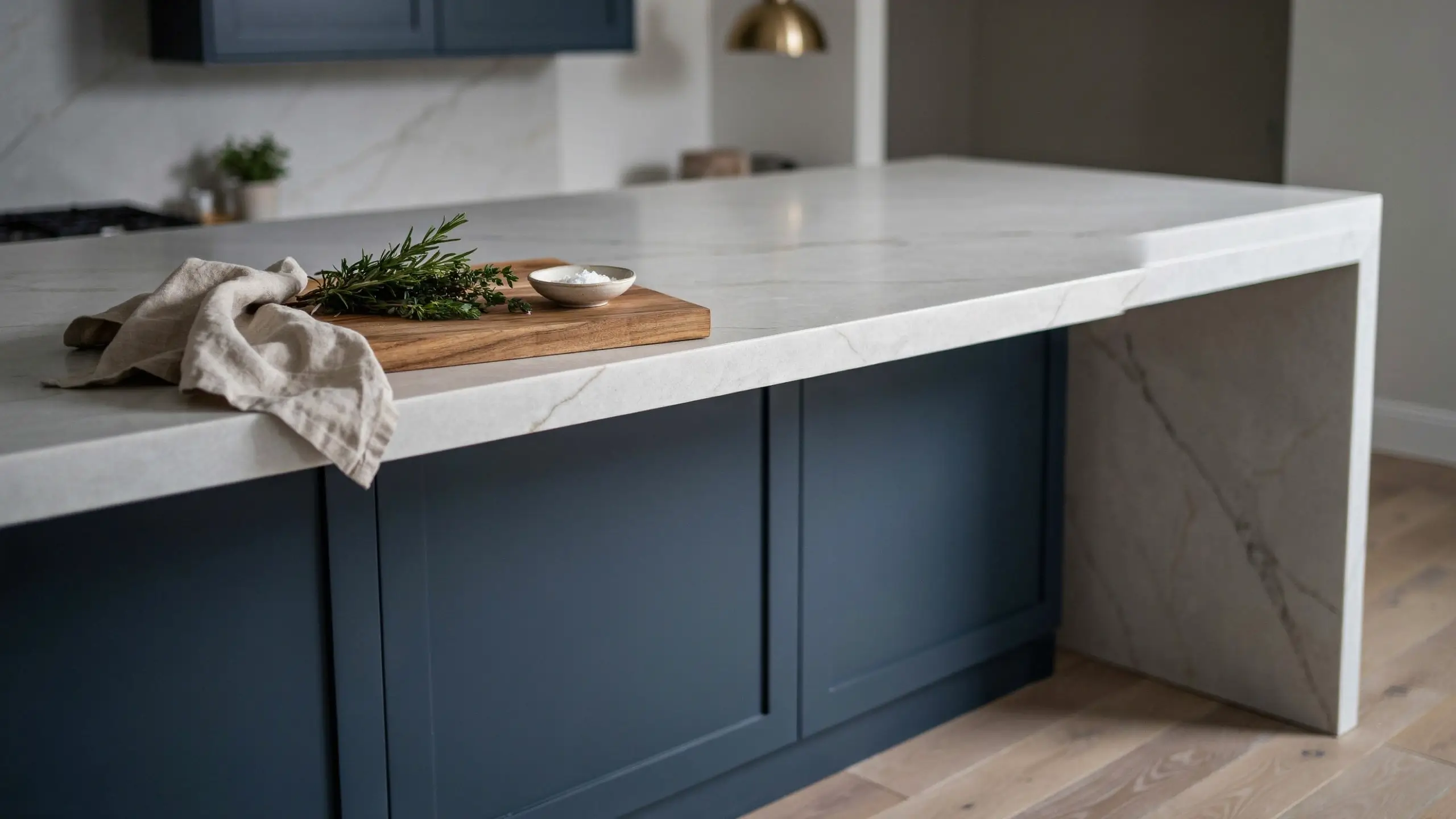 White marble waterfall countertop on a dark blue shaker kitchen island with a wooden cutting board, fresh rosemary, and sea salt.