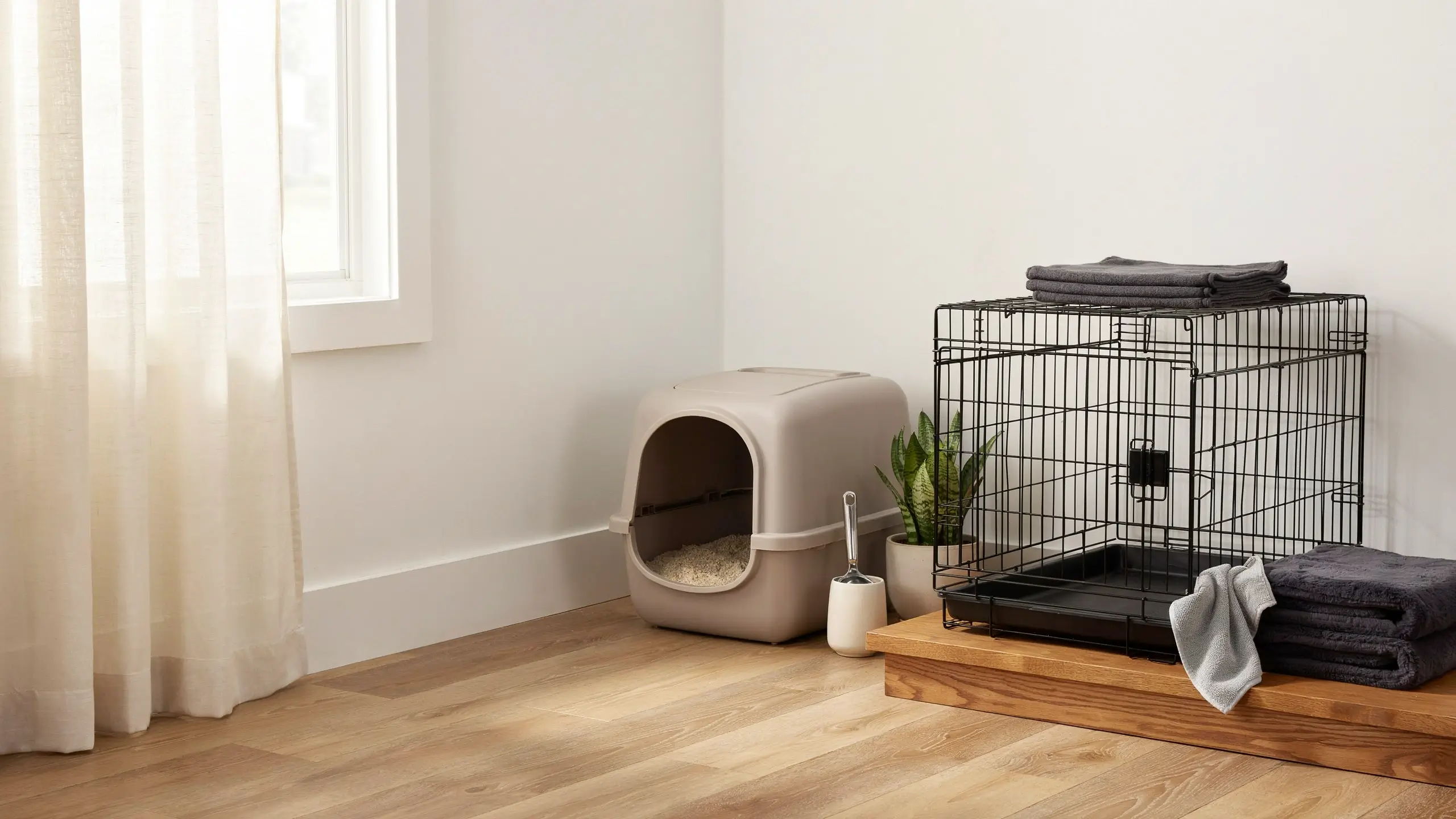 A tidy and aesthetic corner pet organization setup featuring a black wire crate with neatly folded towels on a wooden platform, sitting beside a beige hooded cat litter box with a silver scoop and a small potted snake plant. The functional arrangement sits on a light wood floor next to a window with sheer white curtains, demonstrating a balanced room design.