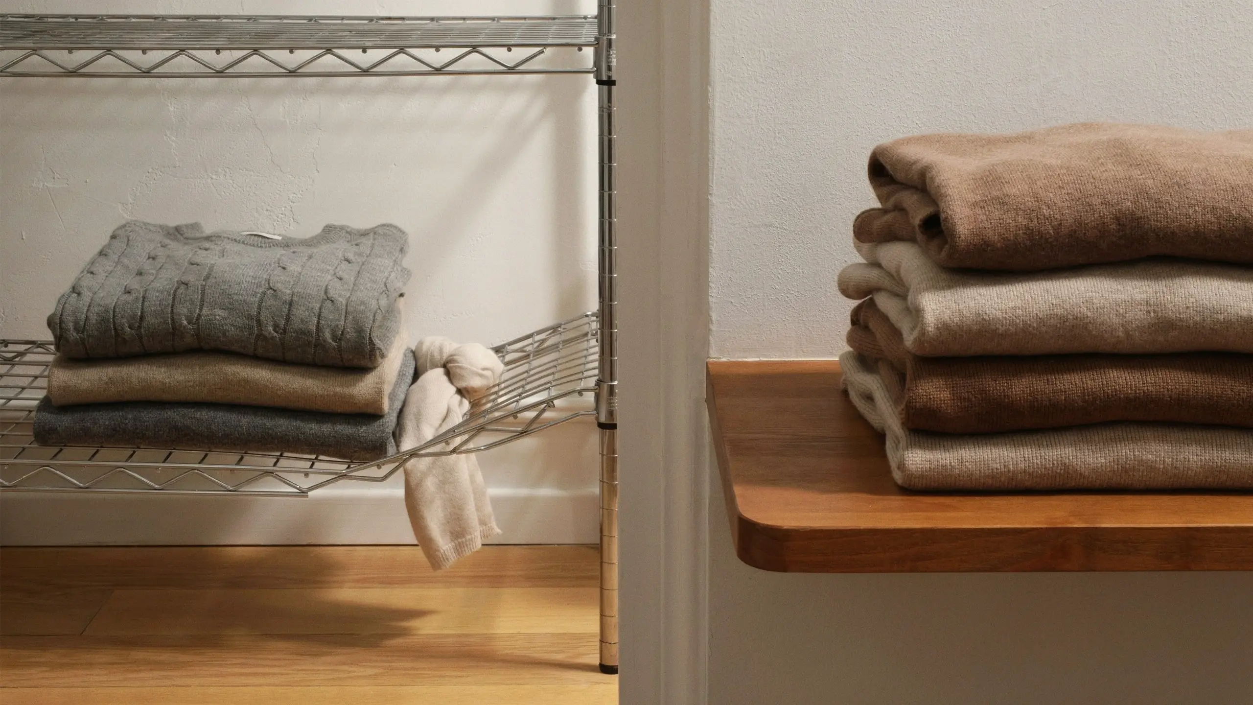 Stack of heavy folded knit sweaters on a metal wire shelf rack with a dangling white garment, next to a neat stack of blankets resting on a smooth wooden storage surface to avoid fabric snagging.